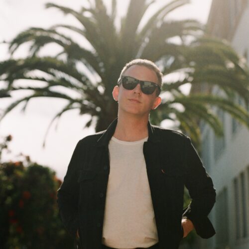 Evan Kennedy, in a square-cropped color photograph, is shown standing in the middle of the frame, cropped from the waist up. He is wearing a black denim jacket, white t-shirt, and black enamel sunglasses. His hands are on his waist and he is looking into the camera. Behind him is a palm tree washed in warm sunlight