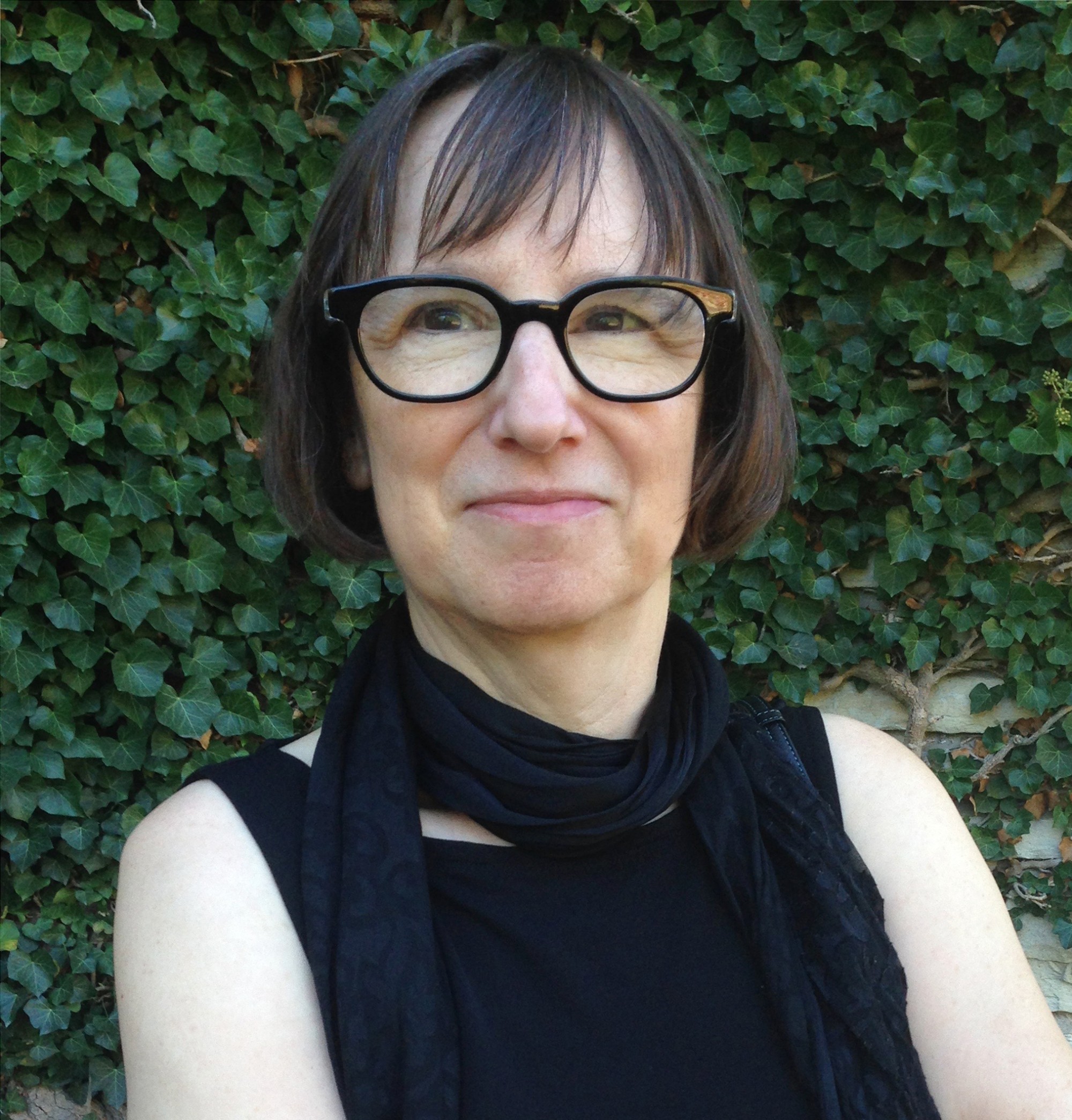 Evelyn is featured in a closely-cropped square portrait, cropped from chest up. She is wearing a black tank top, loose thin black scarf, and black glasses. She is looking just slightly off from the camera and smiling. Behind her is a wall of ivy.