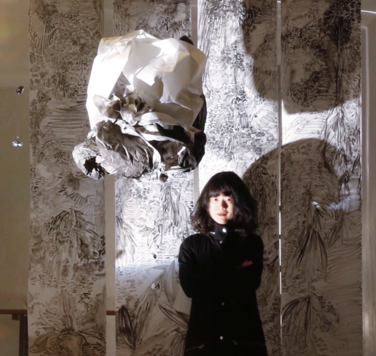 Fei Li is featured center in front of a backdrop, a large drawing of leaves. Fei Li is wearing all black and is standing underneath an orb-like paper sculpture. The lighting in the room is high contrast, with a floodlight on the artist and much of the photograph shadowed or dimly-lit.