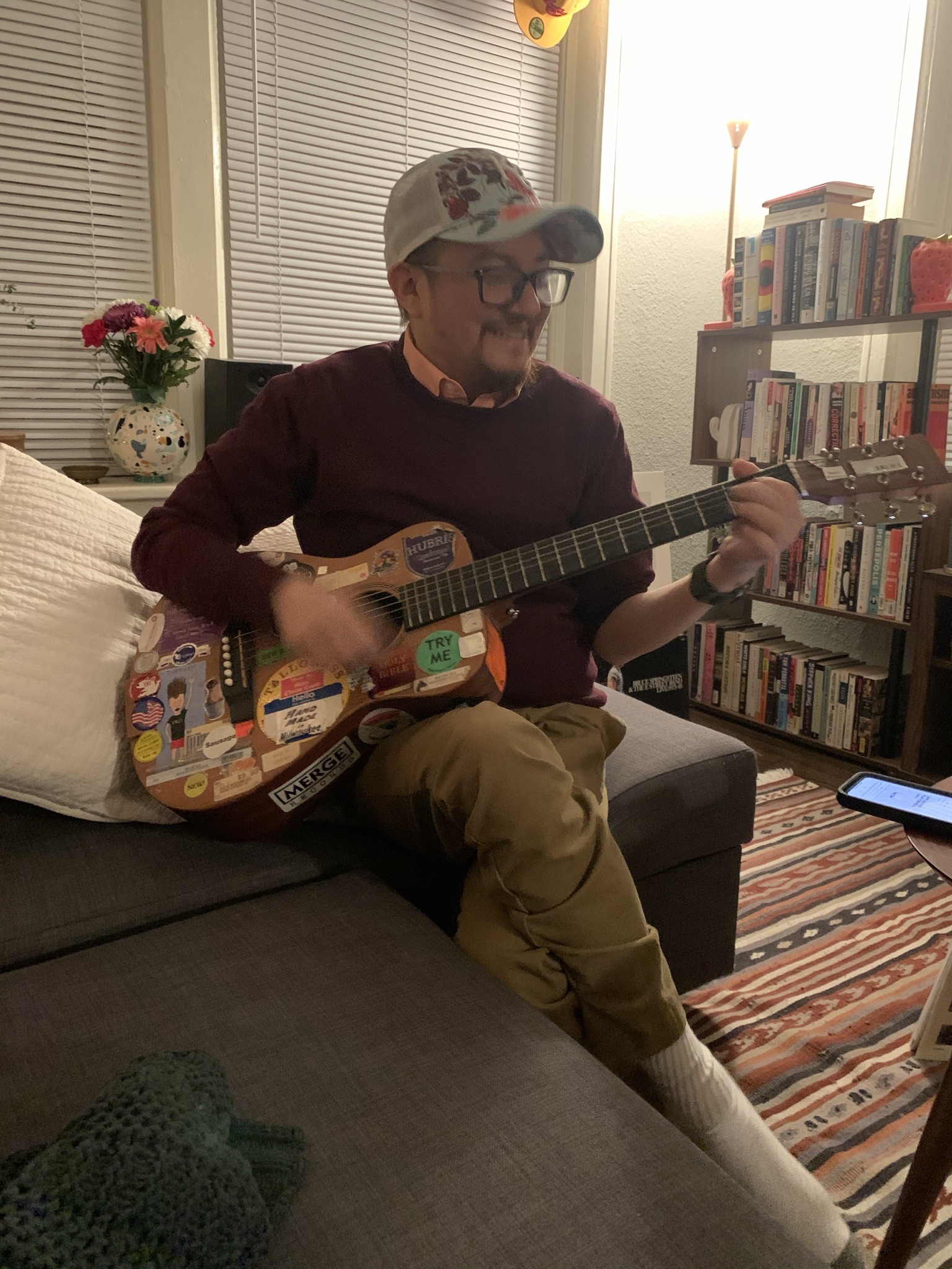 Franklin is seated on the end of a sectional couch, cross-legged, playing a guitar with a lot of stickers on it He is wearing khakis, a red sweater with a button down underneath, and a blue and floral trucker cap. He is looking at the frets of the guitar and smiling. Behind is a bookshelf, lamp, flowers in a vase, and shuttered windows.