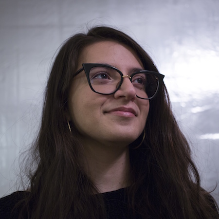 A woman with long brown hair stands in front of a silvery reflective wall. She wears black glasses and a black shirt. She looks towards the top-right corner of the frame and gently smiles.