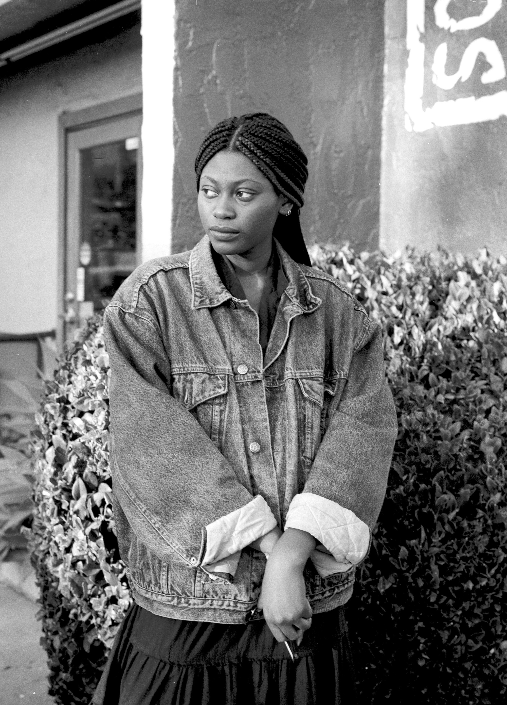 Funto Omojola stands in front of a shrub with hands clasped together, wearing a denim jacket and looking off to the side.