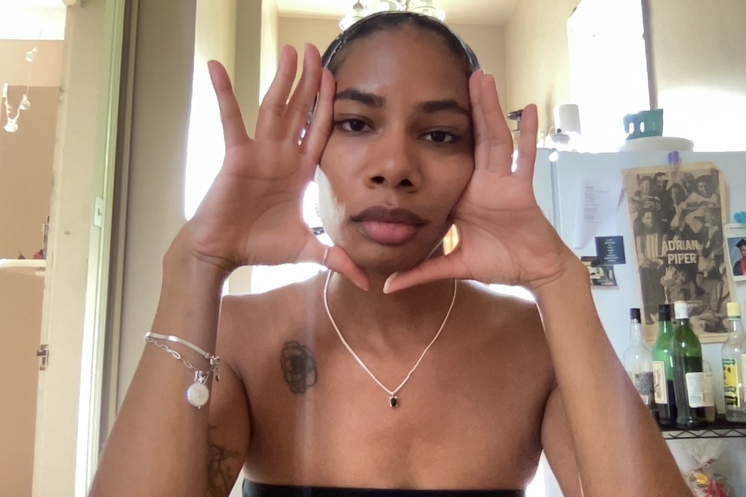 Gabrielle Octavia Rucker sits facing the camera in a black tube top, her hands framing her face, palms faced forward. An Adrian Piper poster is visible in the background.