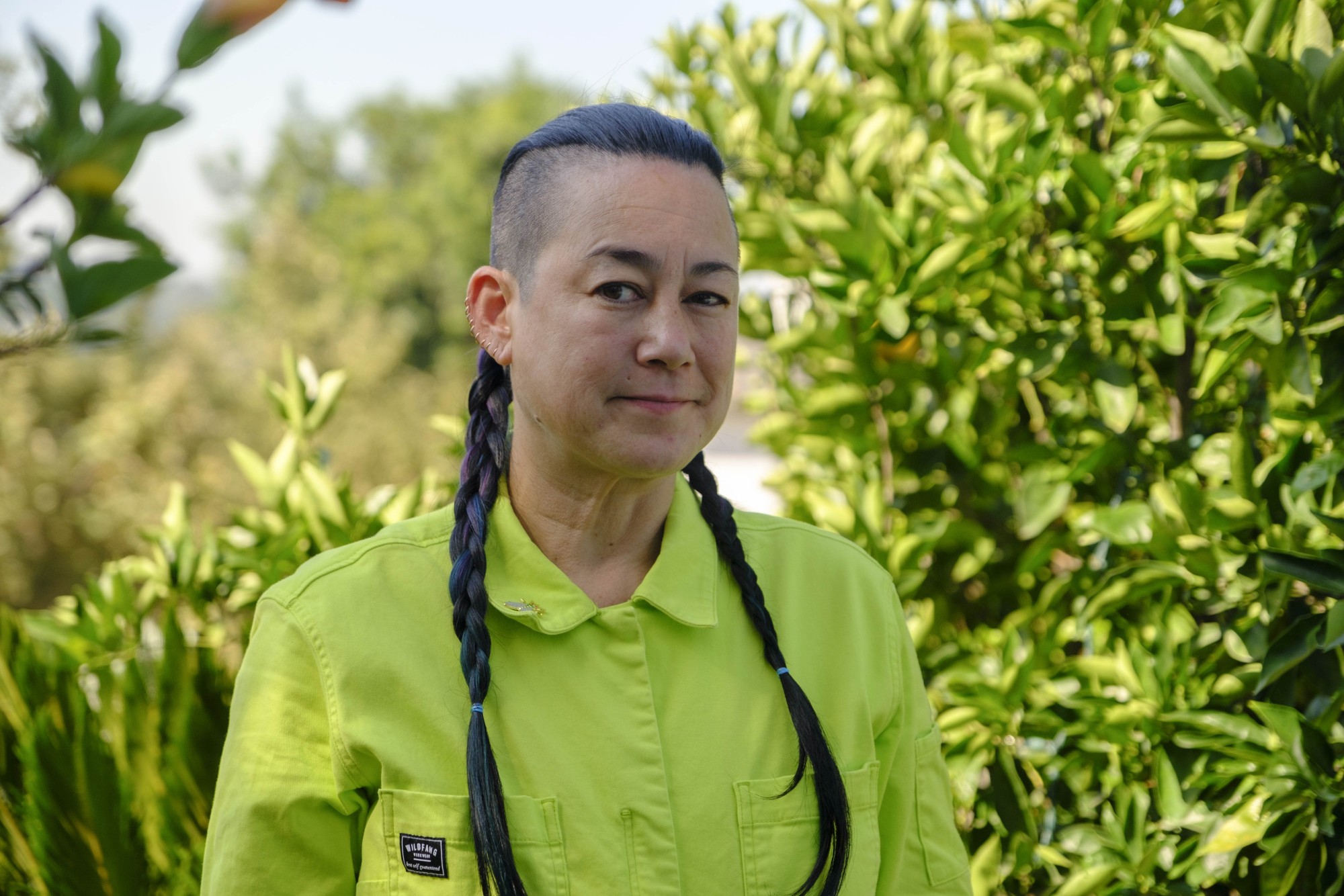 Genevieve Erin O’Brien wears a green button-up shirt, standing outside in front of many green plants. Their hair is shaved on the sides, with two long braids hanging over their shoulders