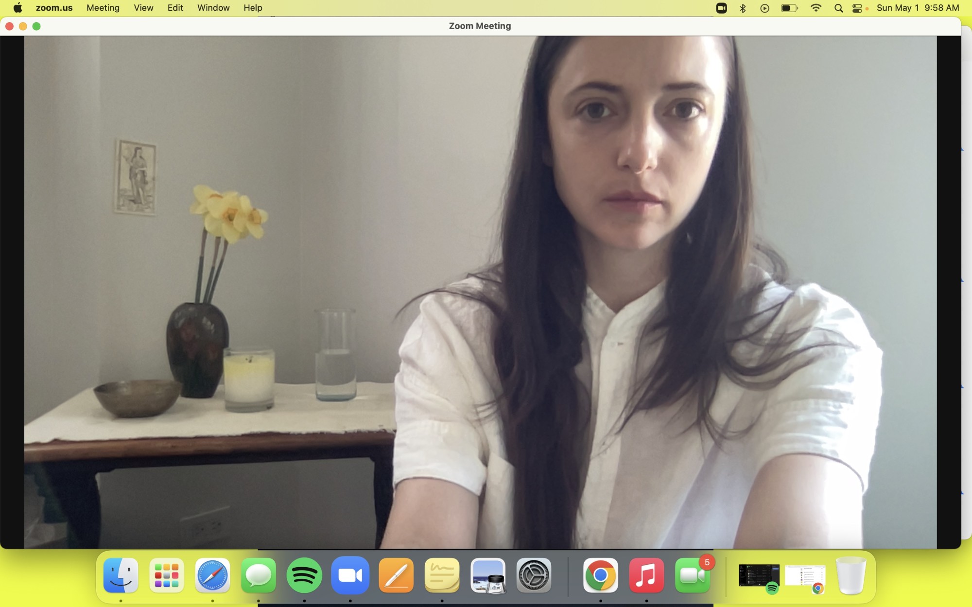 Georgia, a white woman with long brown hair, age 35, is taking a screen shot of herself while using Zoom on her computer.  Below the frame of zoom are icons showing apps that are open. Georgia wears a linen white button up shirt and is looking directly at the camera. Behind her is a table covered in a white cloth. On it sits a candle, a glass of water, a metal bowl and a vase with three daffodils.