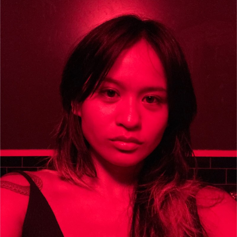 Gia, in a square-cropped photo cropped tightly around the face and from the chest up, looks directly into the camera. The photo is cast in a red light.