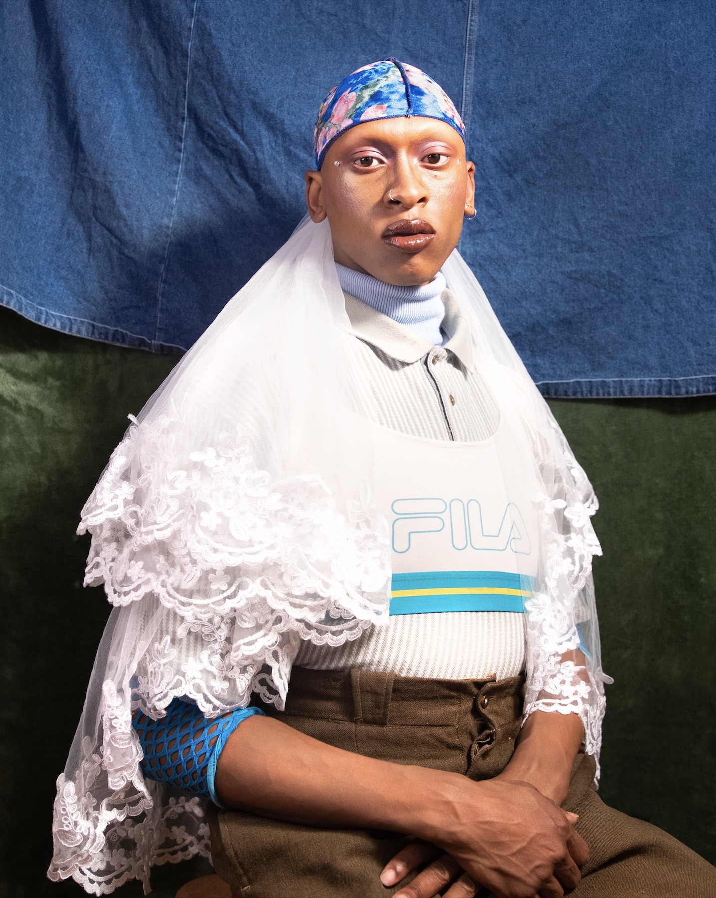 Golden is seated with their hands crossed in their lap in front of a denim and canvas backdrop. They are wearing a blue floral durag with a white lace veil pulled back.