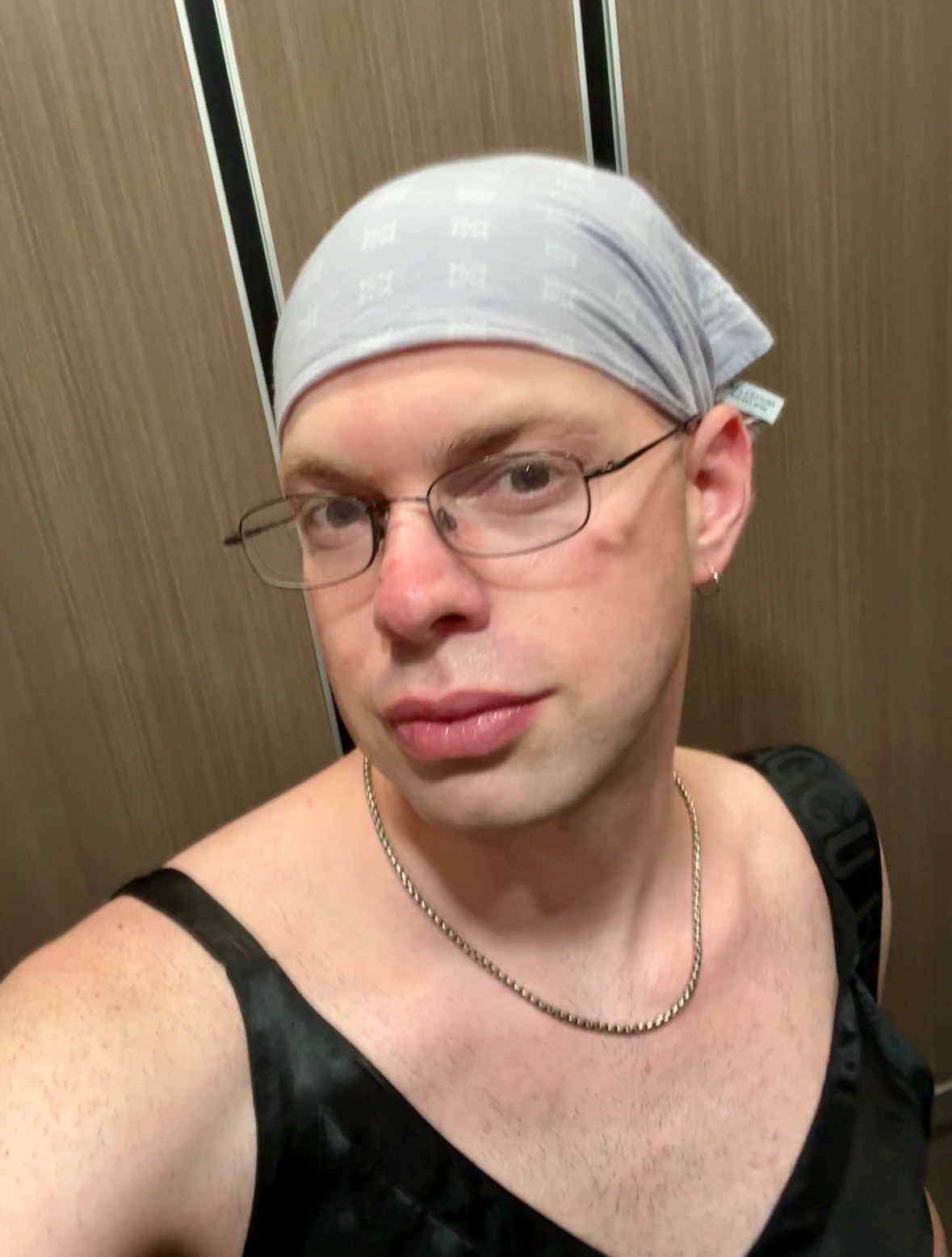 Grace, wearing a black sleeveless top, glasses, and a grey bandana, poses before a wood-paneled backdrop.