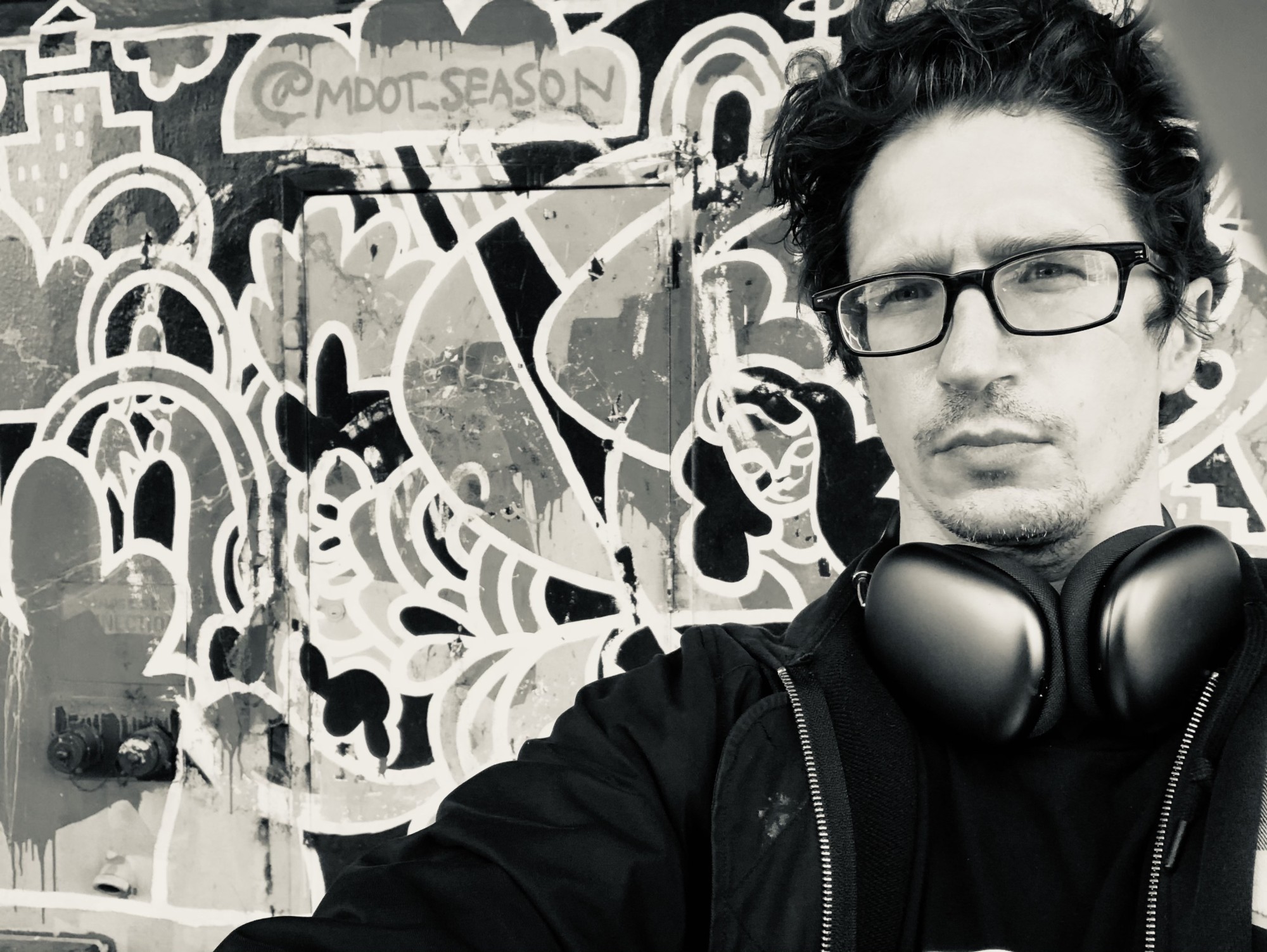 In this black and white photo, Gregory Grube stands in front of graffiti, with large headphones hanging around his neck