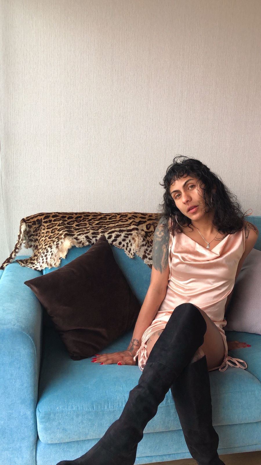 Gretel Warmicha is sitting on a blue couch with a leopard print throw behind them, wearing black boots and a peach slip dress