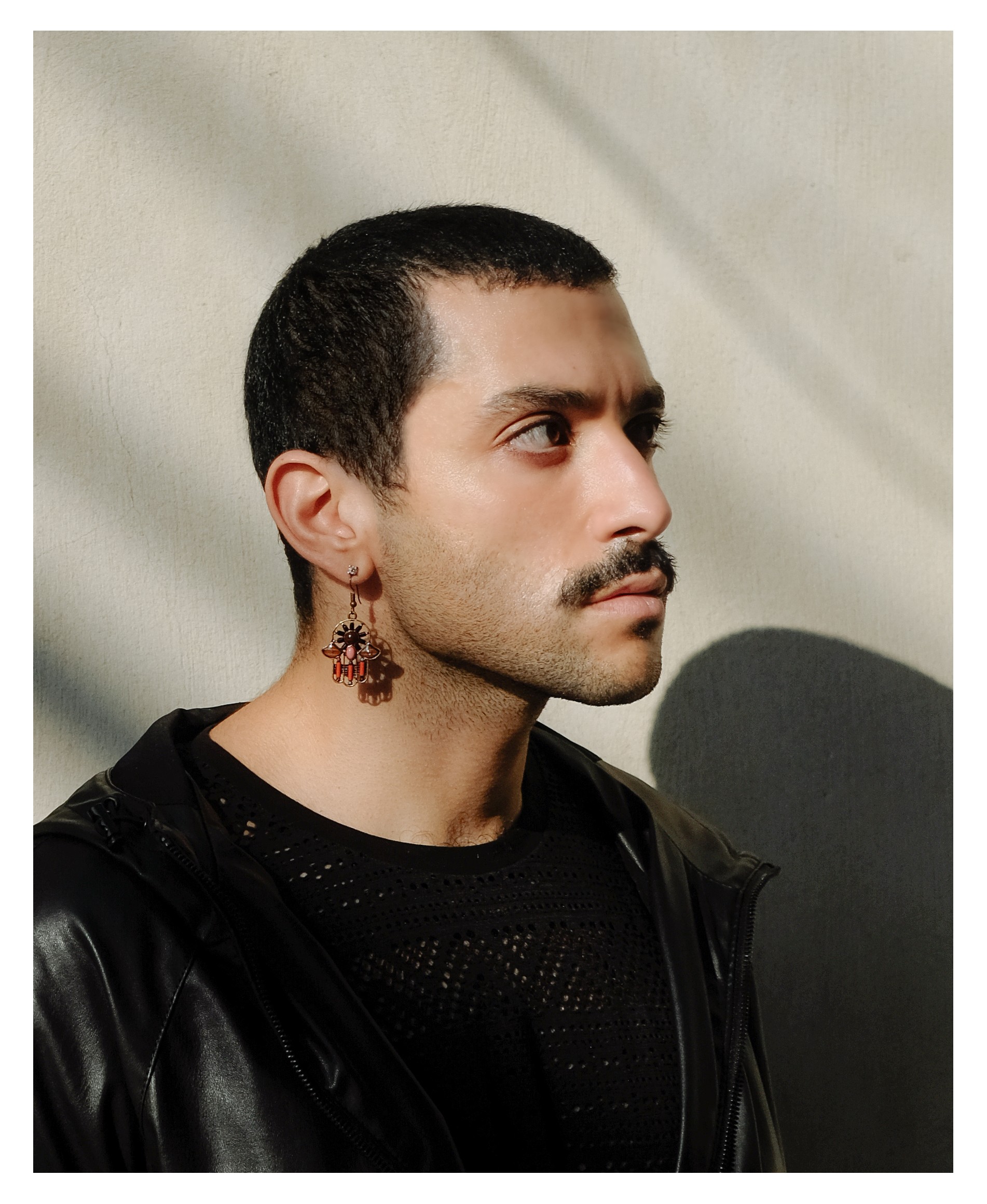 Hamed is featured in a portrait-formatted photograph cropped from the chest up. They are wearing a black t-shirt and a black light jacket. The portrait is in three-quarters, they are facing the right of the frame looking out. They are wearing a dangly earring with a large hamza. Behind is a white wall and bright, angular light.