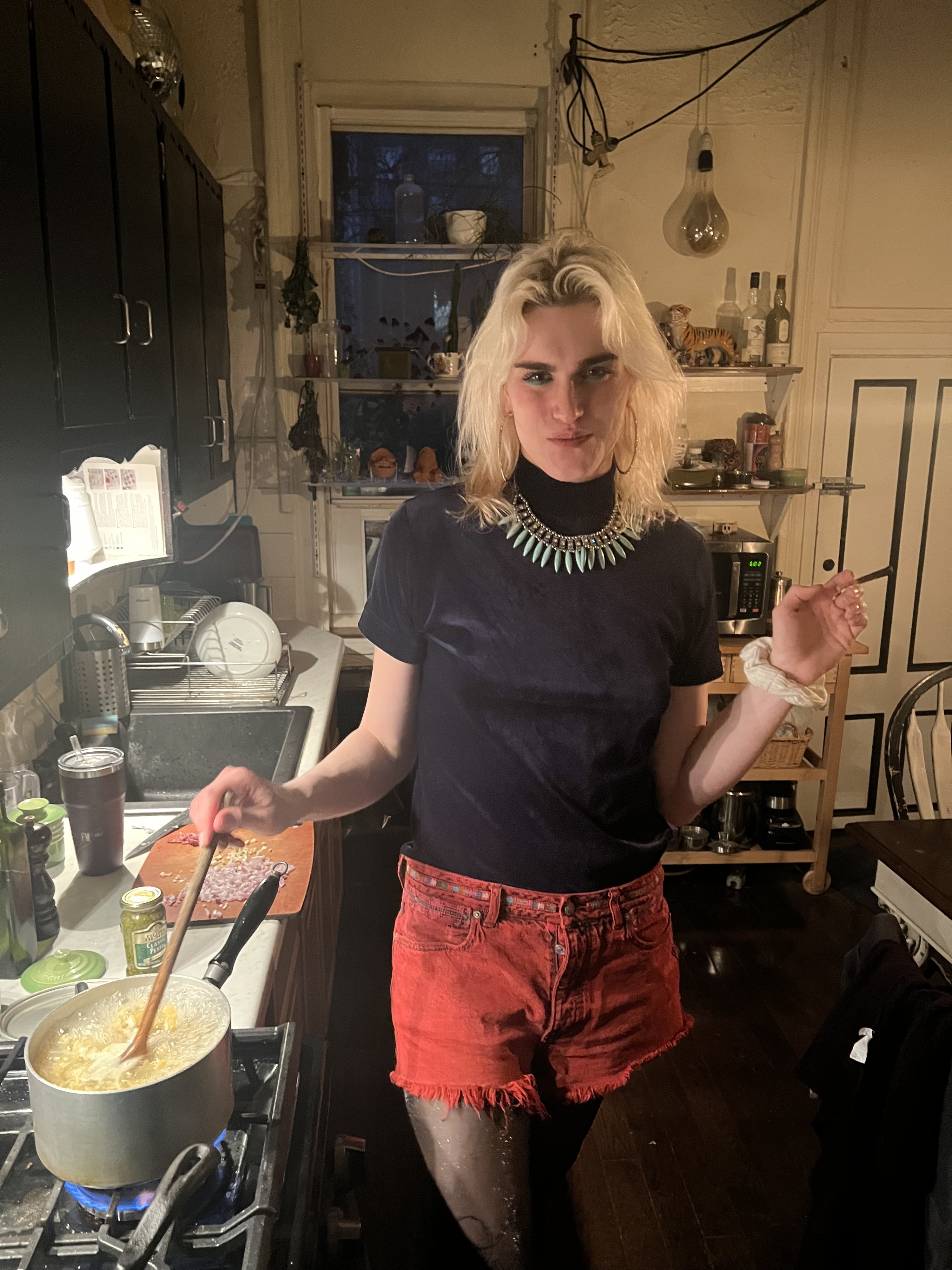 Heather Denton stands at stove in a kitchen, stirring a pot with one hand and smoking with the other. She is looking at the camera and smiling