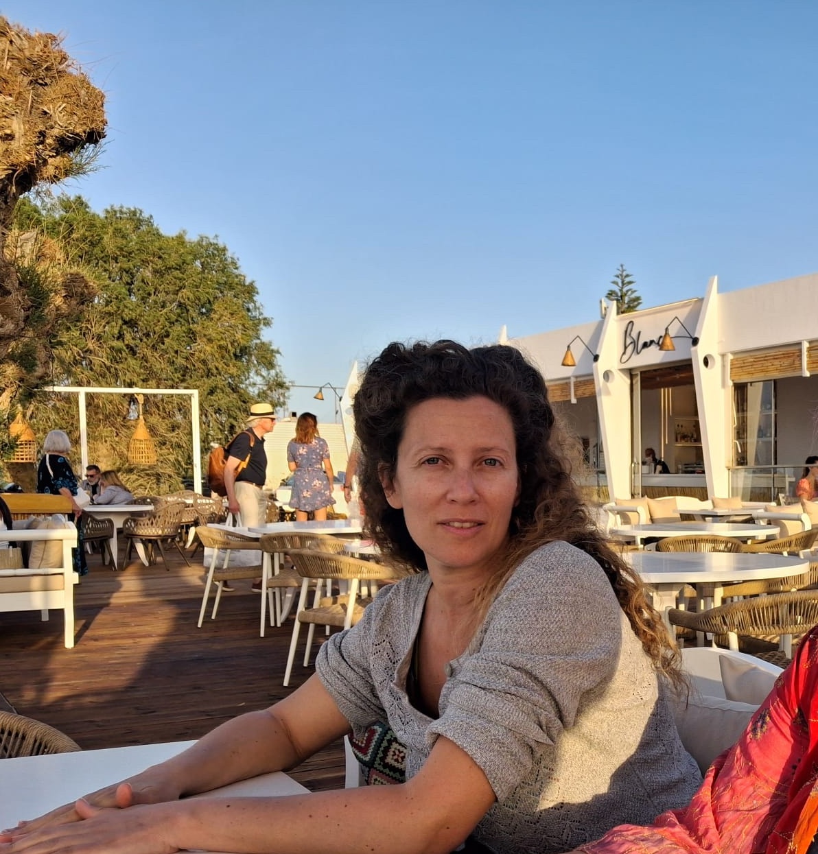 A photograph of Helen Dimos, seated outside at a cafe, looking with a half-smile at the camera head turned. Tamarisk trees, other people standing and seated, and blue sky in the background.