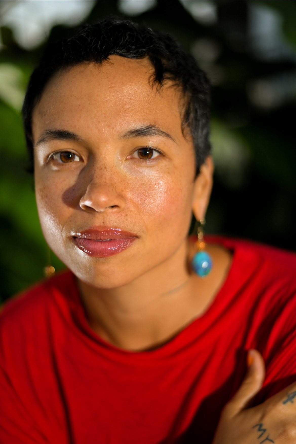 Holland Andrews, a mixed Black person with short black hair wearing a red shirt and looking into the camera and a blurred background of green leaves and dark shadows.