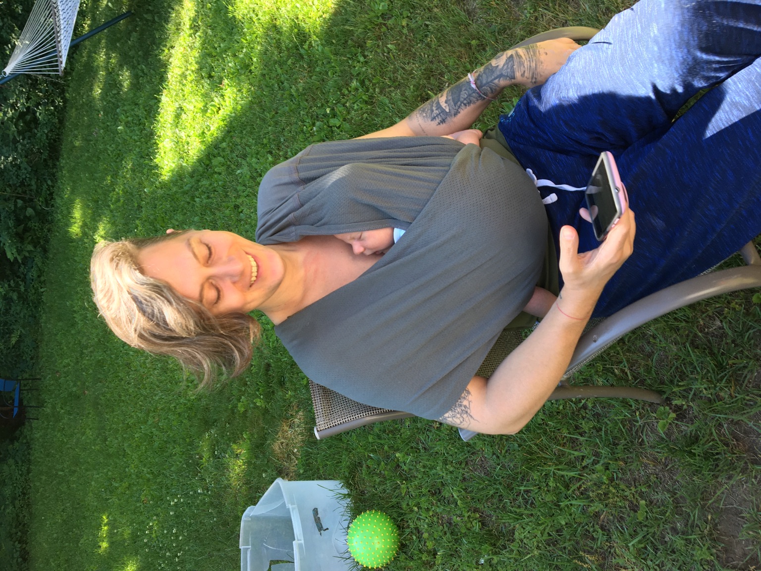 A white trans woman with tattooed arms sits in a sunny, green back yard with a sleeping baby face peeking out of a sling on her chest. She is smiling but avoiding eye contact with the camera by looking at a phone in her hand. At least one side of her head is buzzed; the rest of her hair is brunette and chin length.