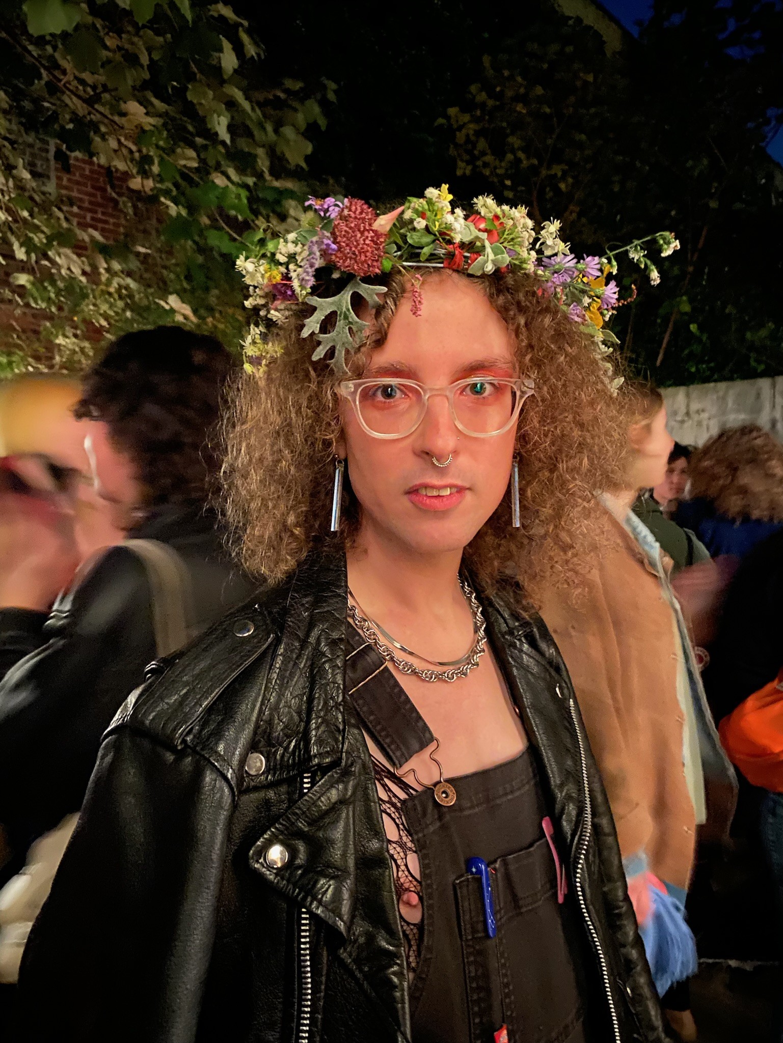 imogen is featured in a portrait-oriented picture cropped from mid-waist up. They are wearing black overalls, a black leather jacket, dangly earrings, large clear enamel-framed glasses, and a flower crown. Behind appears to be an outdoor party. Trees and blurs of people are visible in the background.
