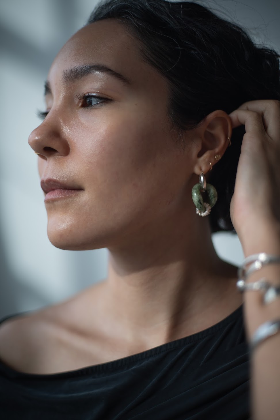 A photograph of India staring off to the right, gently pushing her short black hair behind her left ear. She is wearing gold, silver, and stone-based earrings, silver bangles on her left arm, and a charcoal dress.