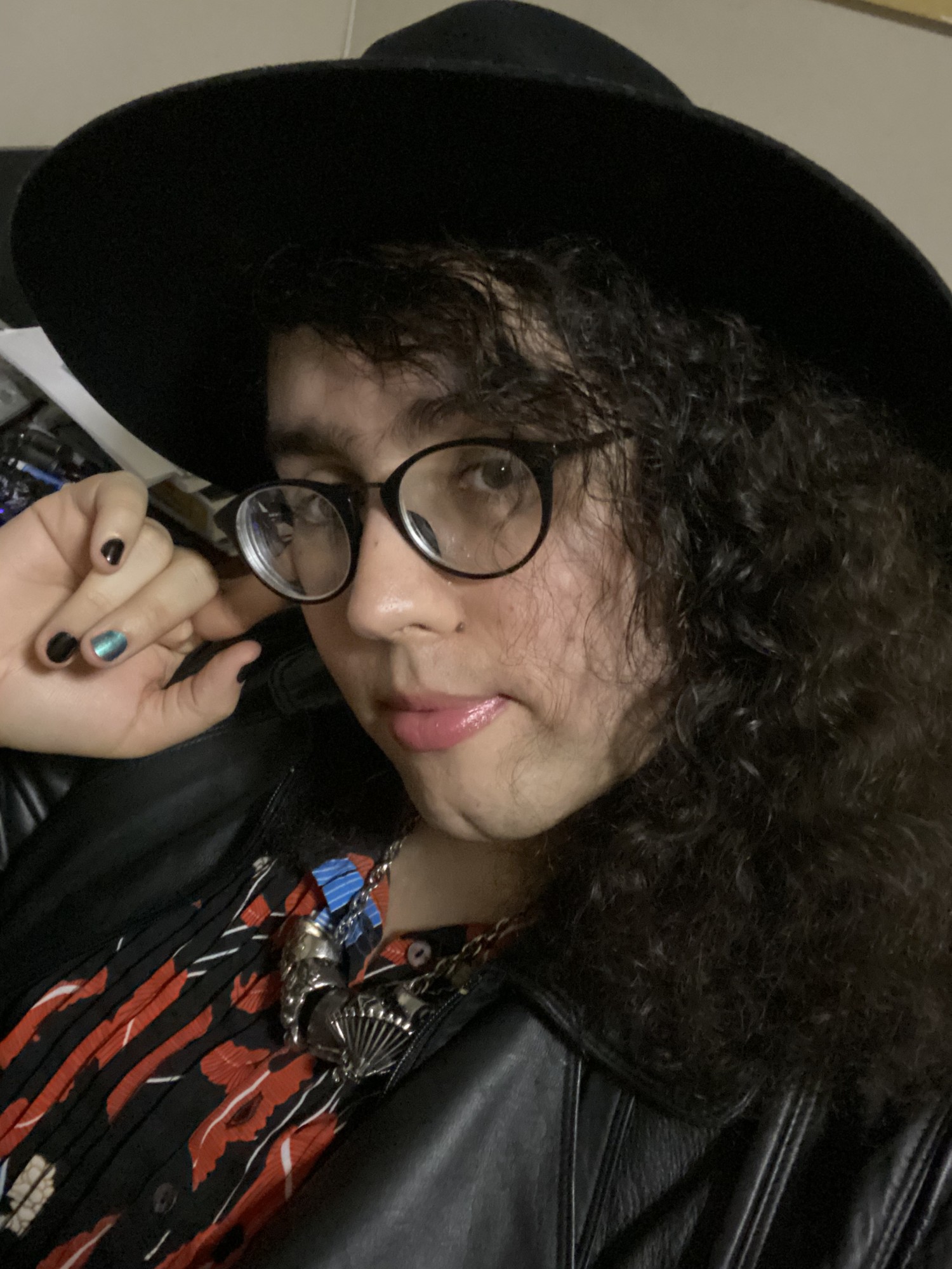 In this color portrait, Isa Guzman looks at the camera, her cheek resting on her hand, her nails painted a dark metallic green. Guzman is wearing a leather jacket, black glasses, and a large-brimmed black hat