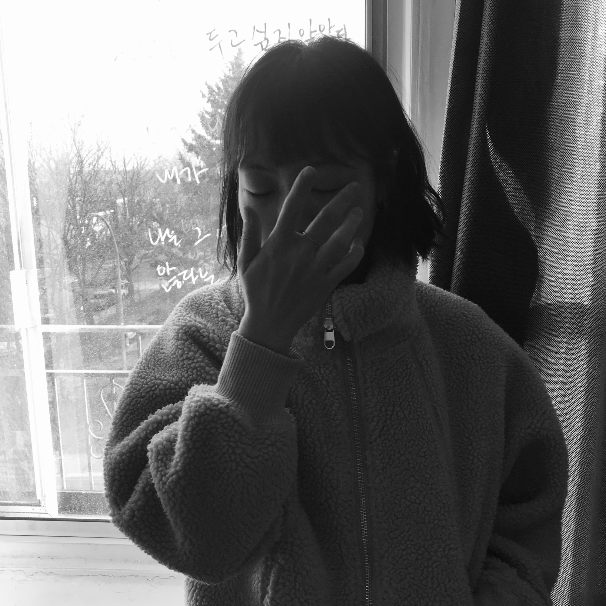 this is a square black-and-white photo of Ivetta. She is centered in the picture, wearing a wooly fleece jacket, and is covering her face with her right hand. She stands in front of a bright window.
