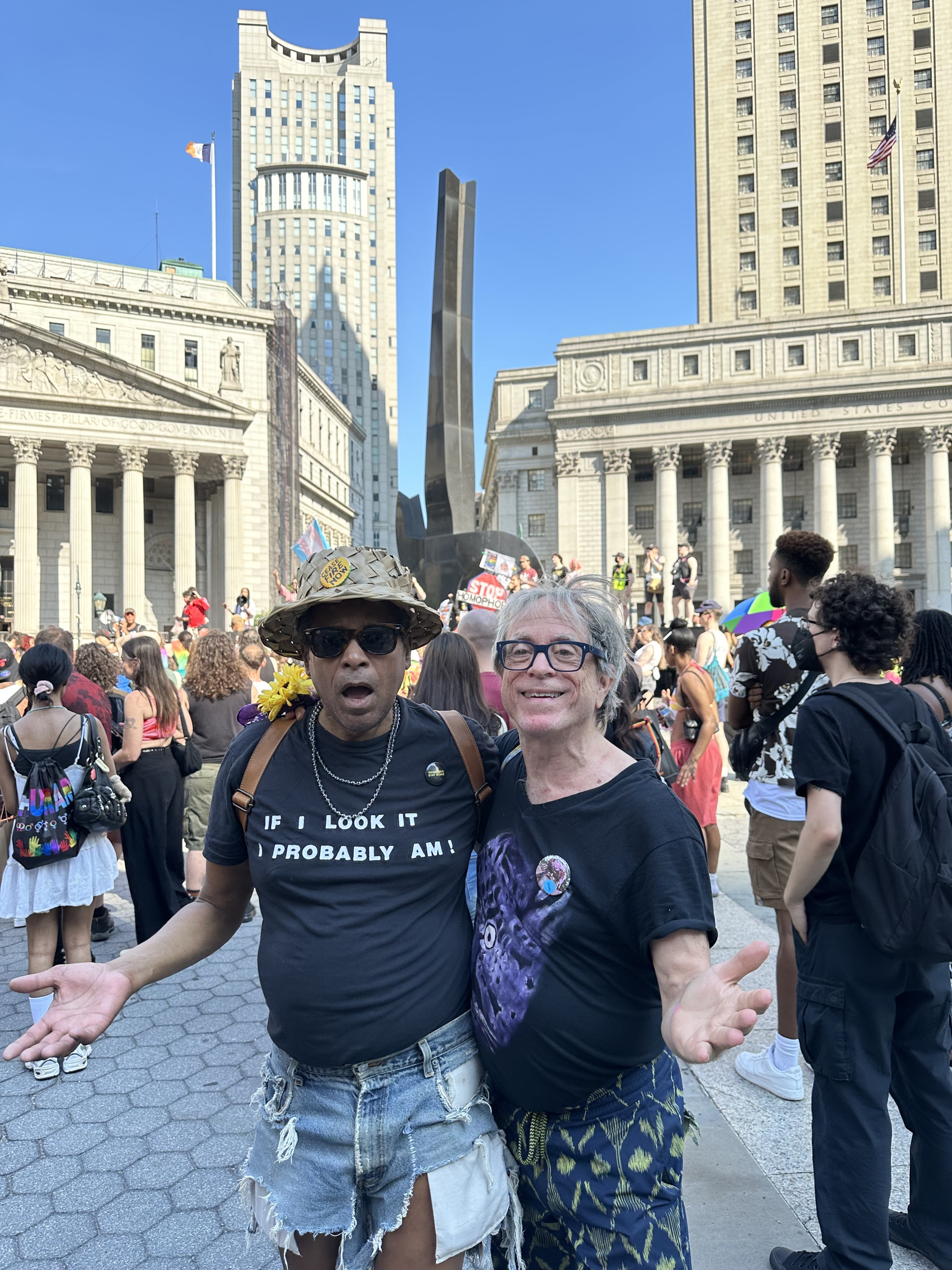 At Foley Square ending the Reclaim Pride march Jack is in the left foreground wearing sunglasses and a straw pork pie style hat with a button saying "Cease Fire Now" in the middle of the front wall of the crown. His black t-shirt reads in simple white block letters: "If I Look It I Probably Am!" Peter is on the right wearing black framed eyeglasses. Peter's shirt is also black but more colorful with a fantasmagorical image of a blurry creature that could be a tiger or a dragon. They are embracing so that their joining arms are concealed behind each other's back while each free arm is outstretched to the side, Jack's in a enquiring gesture of delighted surprise, Peter's in charmed welcoming. They exude an asymmetrical balance signaling the impulse to embrace of the viewer. Jack's mouth is softly agape, Peter has a toothy, but relaxed smile. In the background Queers attend the speakers in a curved semi-circle that imitates J&P's stance. The court buildings are sunlit. The signage is illegible from this distance. The white stone neoclassical municipal structures are contrasted by a modernist abstract black steel sculpture whose veriticaly upward rise resembles a giant finger that softly centers the composition dividing the architectural background as it splits J&P in the foreground. Jack is black and Peter is white.