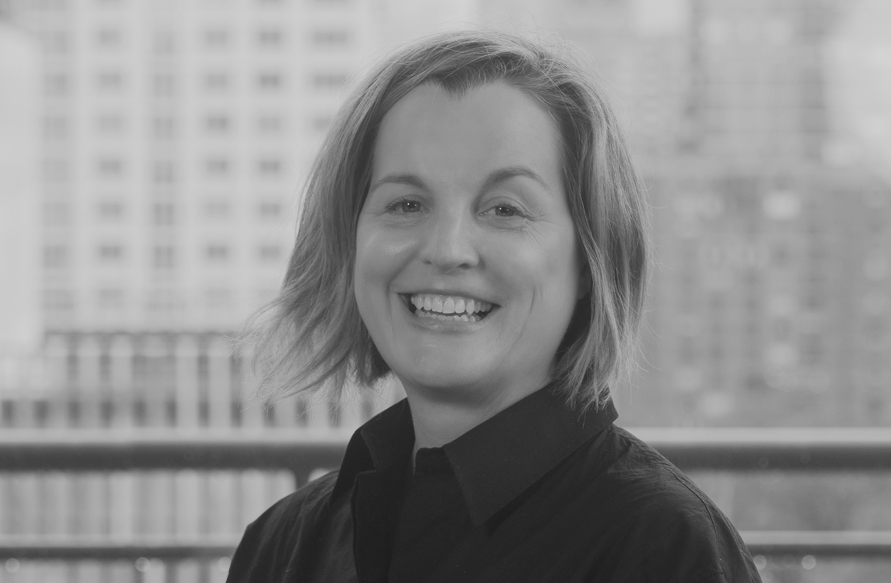 A black-and-white headshot of a white woman with medium blond hair, photographed in front of a window through which buildings around New York's Lincoln Center are visible. She is wearing a black collared shirt and smiling.