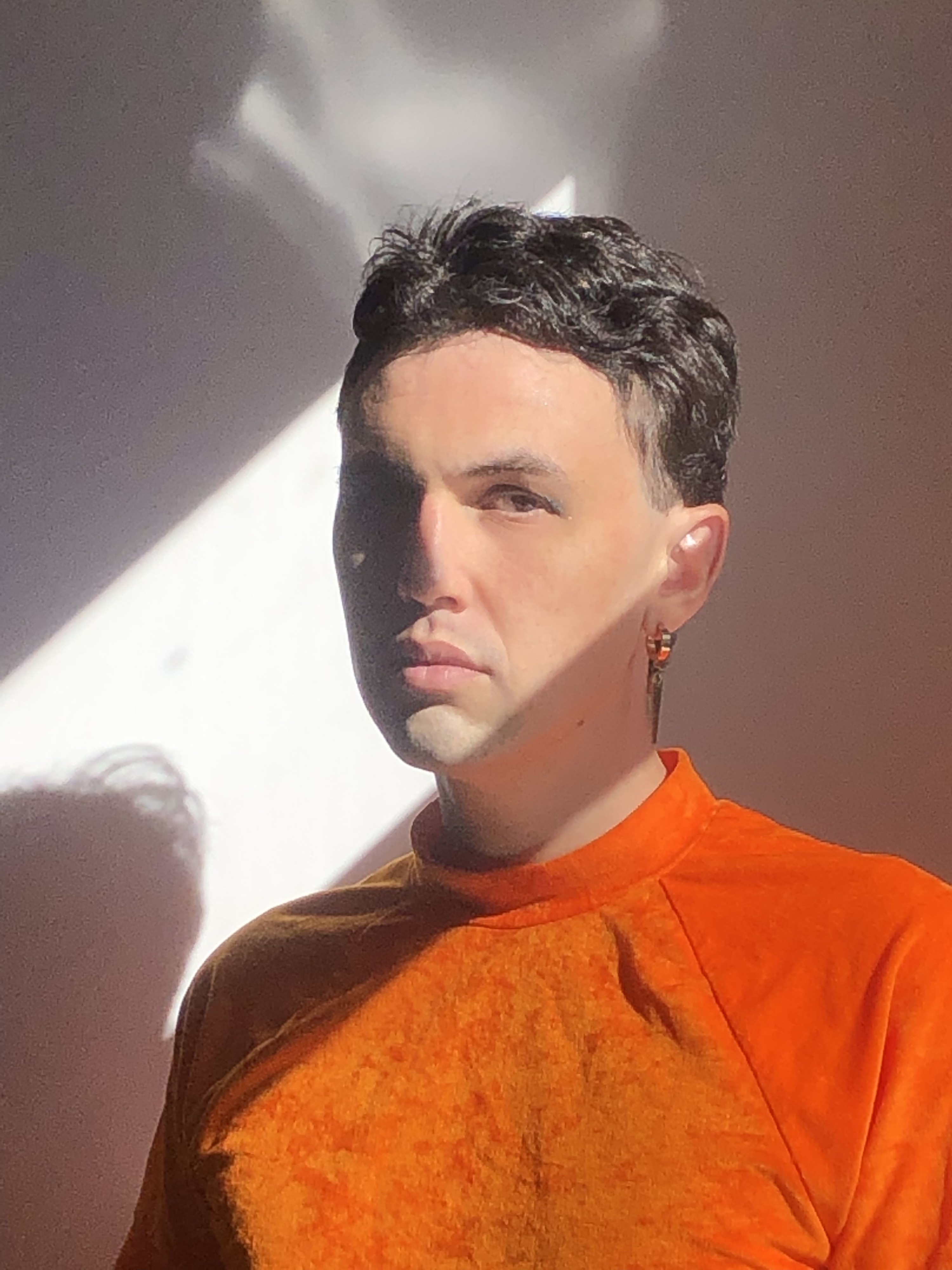 author is standing by a sunny window. they are wearing an orange tee-shirt, an earring, and green eye shadow.