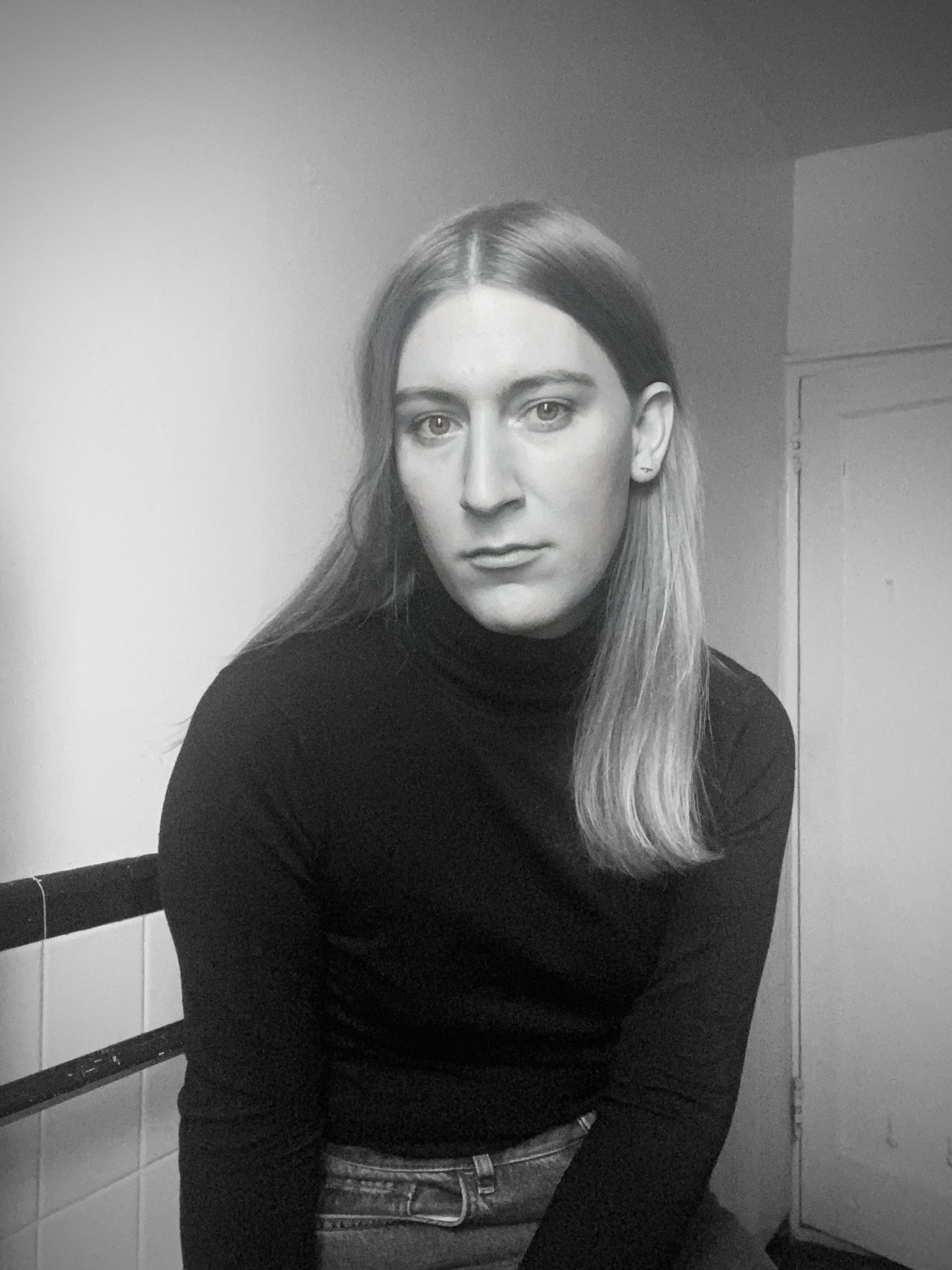 A black and white photo of Jameson Fitzpatrick looking at the camera. Jameson is wearing a black sweater and has long, blonde hair.