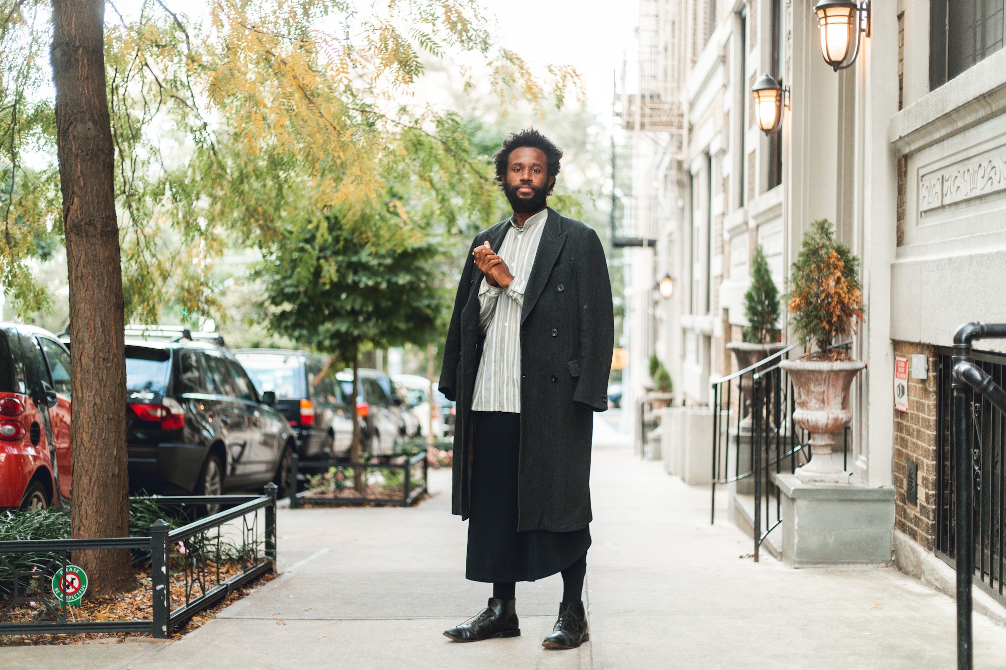 JJJJJerome Ellis stands on the sidewalk between a street tree and building, wearing an overcoat over their shoulders, their hands clasped at their heart, looking into the camera.