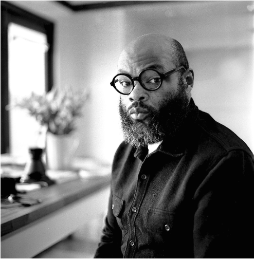 Black and white image of Jerriod, a Black man wearing a dark button up shirt with a white undershirt, round dark glasses frames, sits in a sunlit room. A white vase filled with flowers sits atop a counter in the background, amidst various other objects.