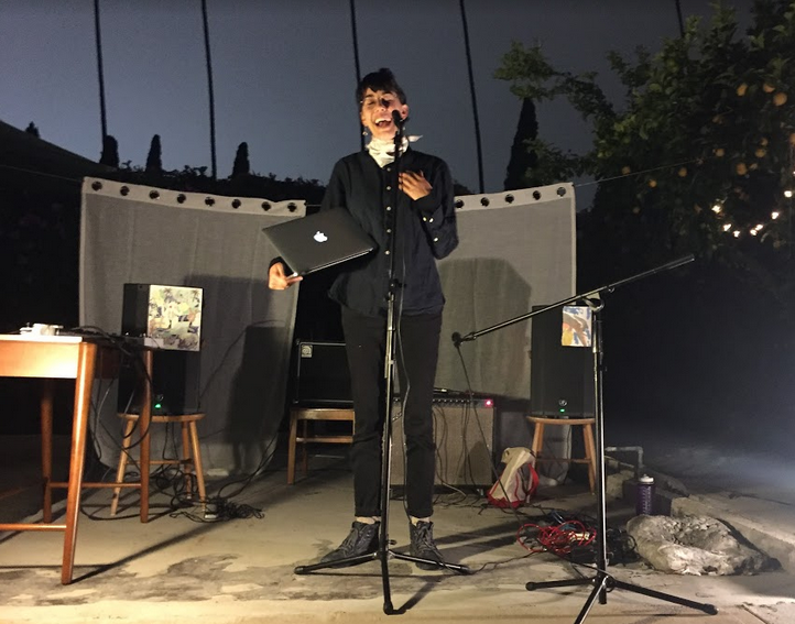 Jhani/J F K Randhawa is giving a reading outdoors in the evening, standing in front of a microphone, dressed all in black, with various equipment behind and around them, as well as two curtains, and a citrus tree visible to the right. Their eyes are closed as they speak, and their hand is at their heart.