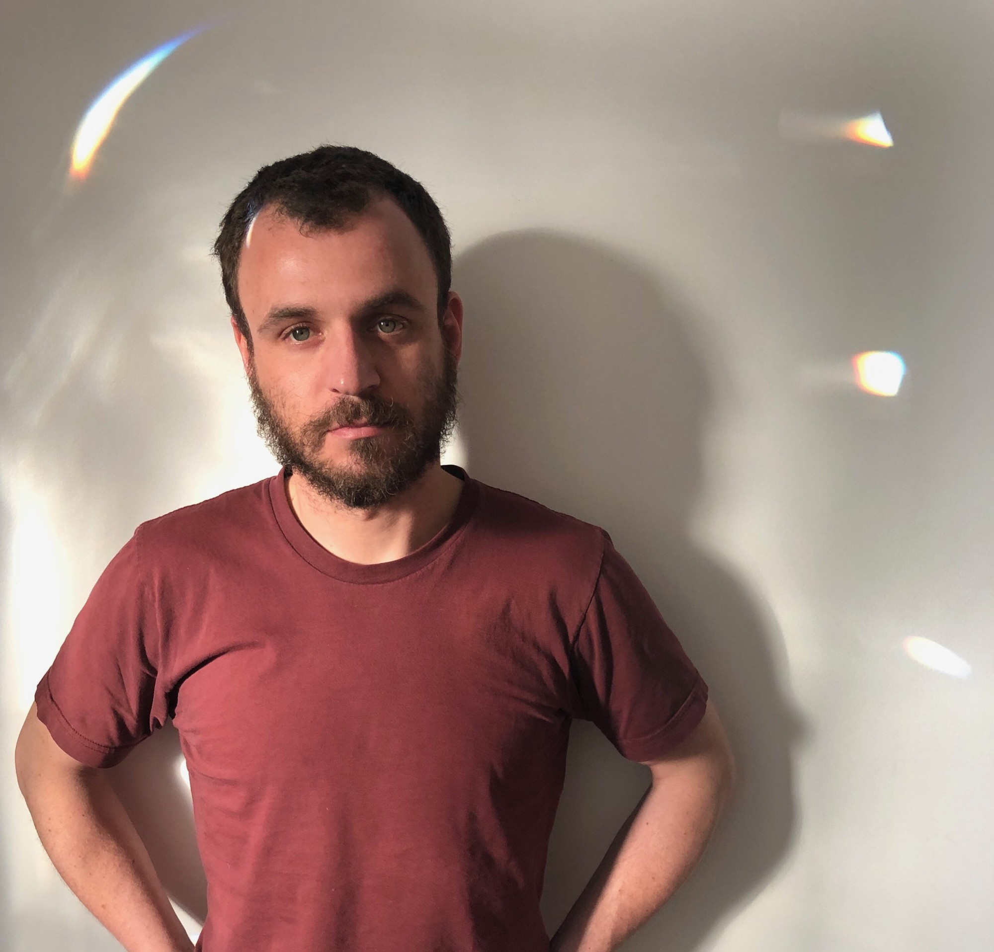 Josef Kaplan stands in a red t-shirt in front of a white wall, dappled with refracted light, looking at the camera