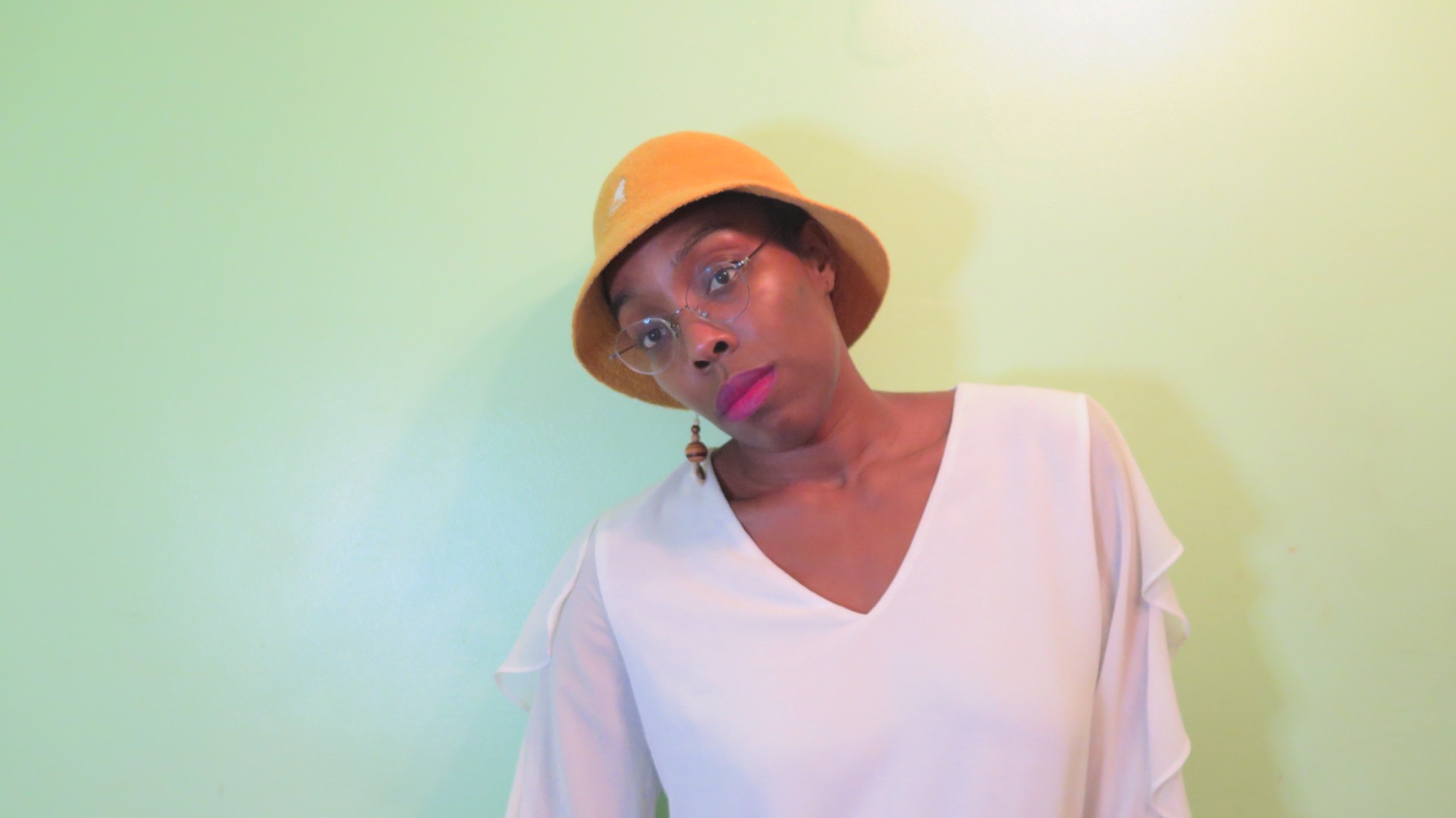Joselia Rebekah Hughes stands in front a pale green wall. She's leaning slightly to the left, wearing a yellow bucket hat and magenta lipstick.