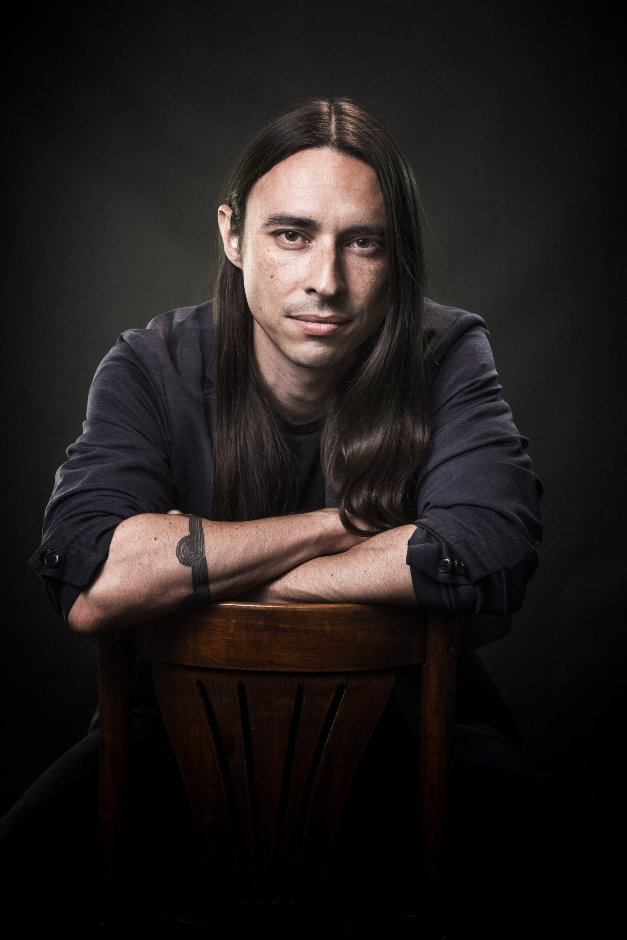 Joseph is featured centered in the photograph in a studio with a black backdrop. The photograph is largely black except for where Joseph is shown, leaning his forearms on a stool, facing the camera and smiling. He is wearing a black long sleeve shirt.