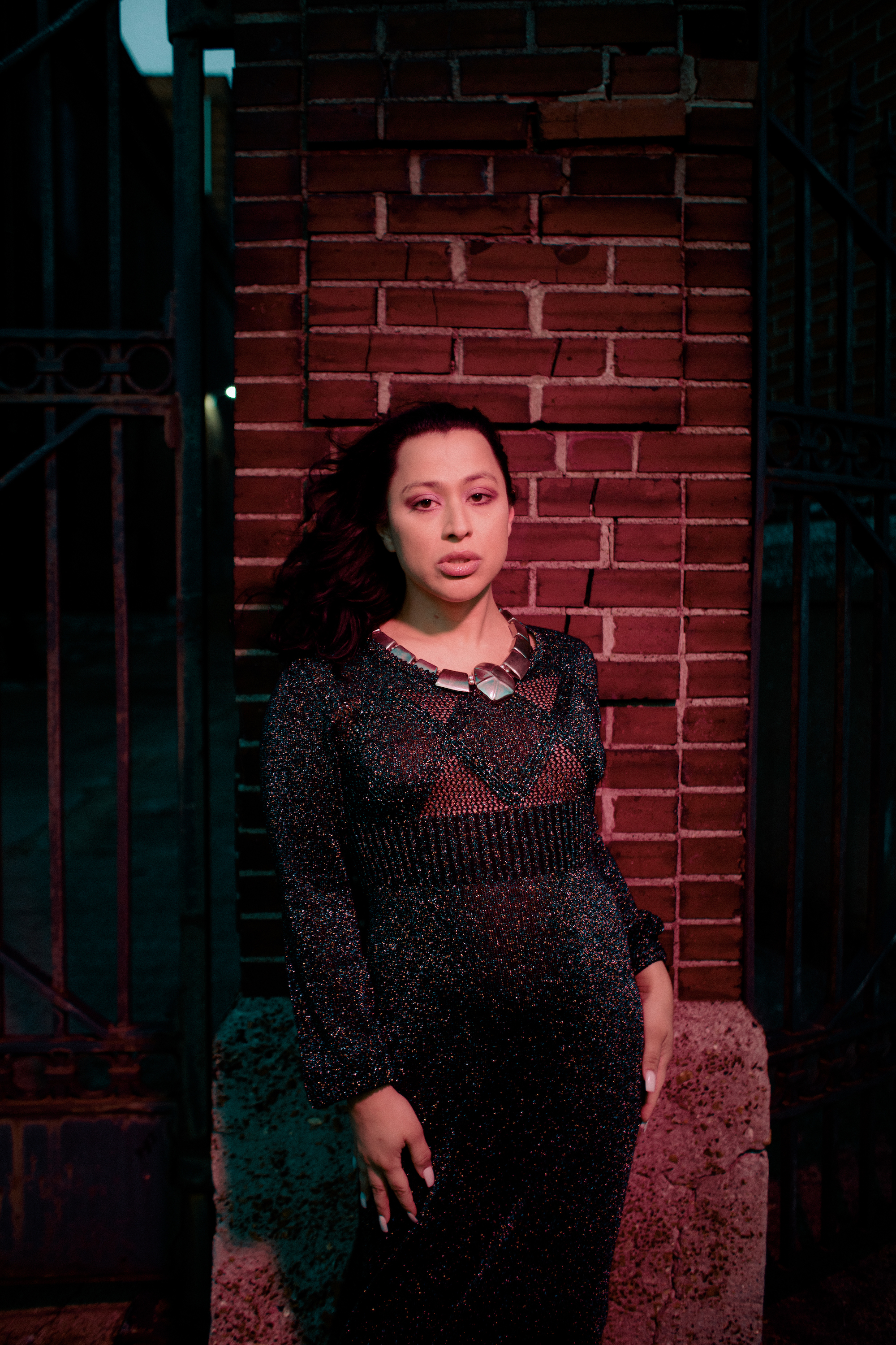 Joss Barton, a light skin latina transsexual woman, stares at the camera. Her hair is black and a few strands are being blown on her face by the wind. She wears a silver round necklace and a dark iridescent knit gown while standing in front of a red brick wall.