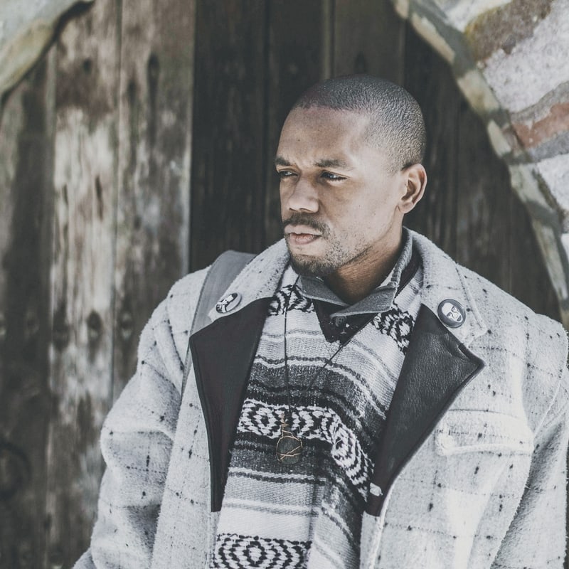 Justin Phillip Reed is standing outside in front of a wood door wearing a gray coat, looking to the left.