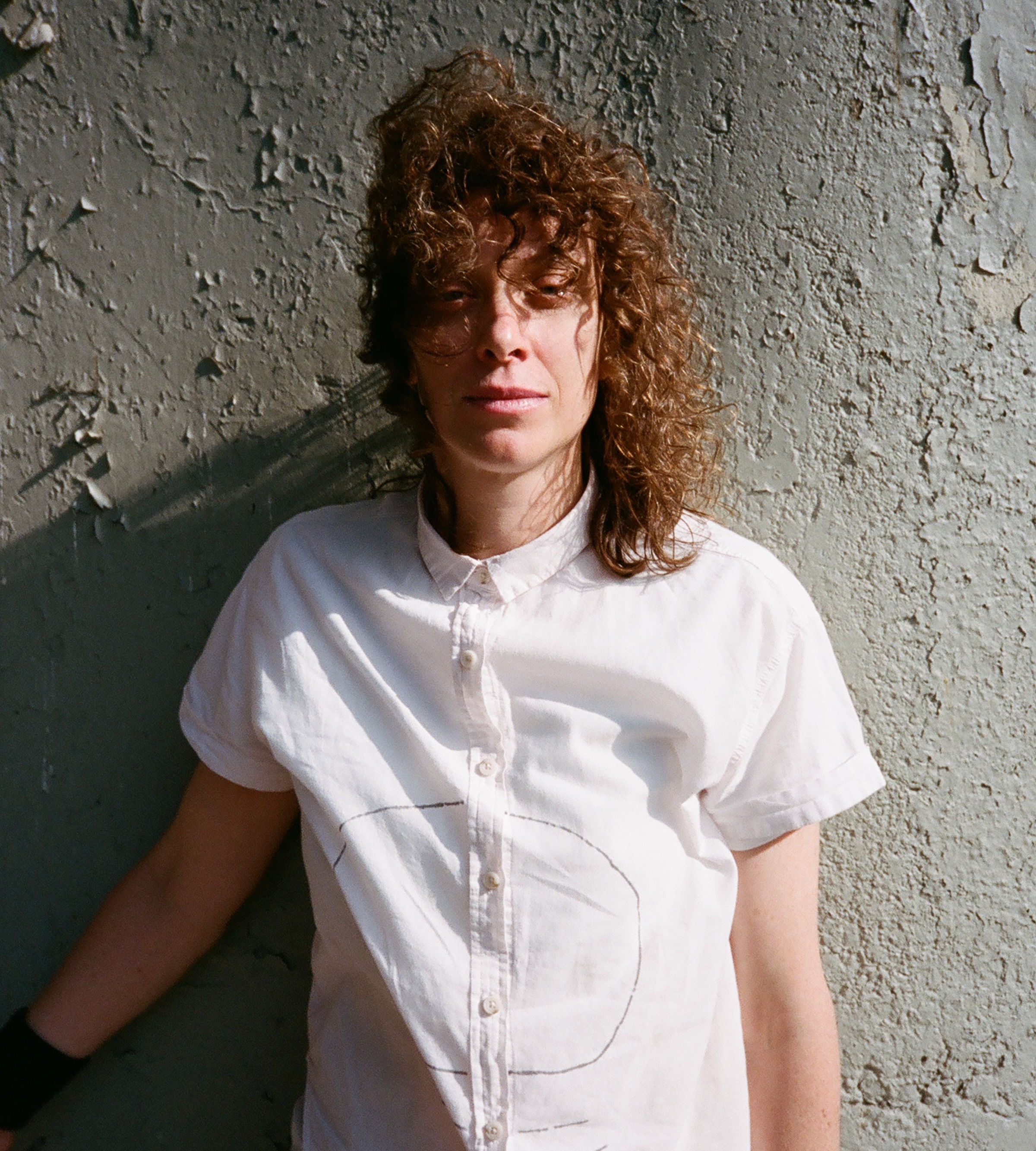 A nonbinary person with brown long curly hair stands against a textured wall outside in sunlight with a light breeze. They are wearing a white cotton short- sleeve button down shirt with a large circle made with a BIC pen prominently displayed on front.