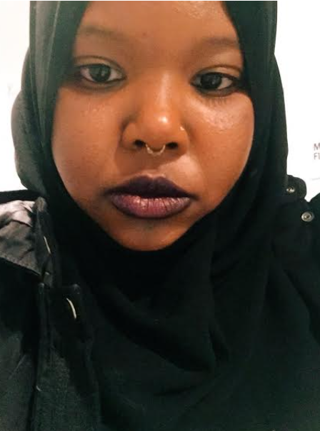 Kemeelah in closely cropped frame wears purple lipstick, a septum piercing, a black jacket, and a hijab.