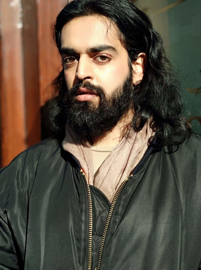 Kashif wears a jacket and looks into the camera, with long hair and a beard