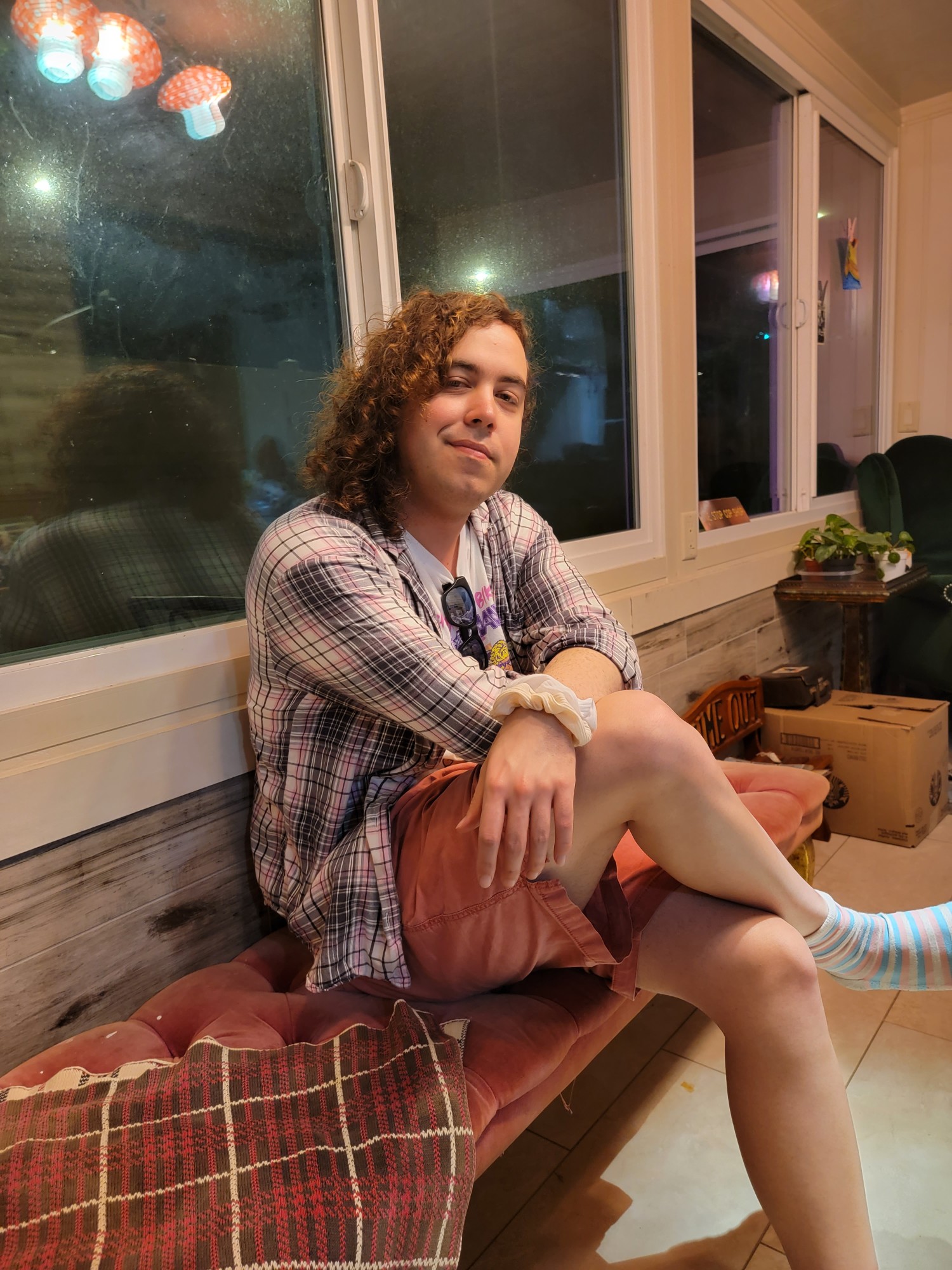 Kass is shown seated cross-legged on a cushioned bench with windows behind her, facing the camera at a three-quarter angle. She is wearing orange shorts, a grey plaid button down, unbuttoned with a white t shirt underneath it. Her brown curly hair is loose over her shoulder, with a white scrunchie on her wrist closest to the camera.