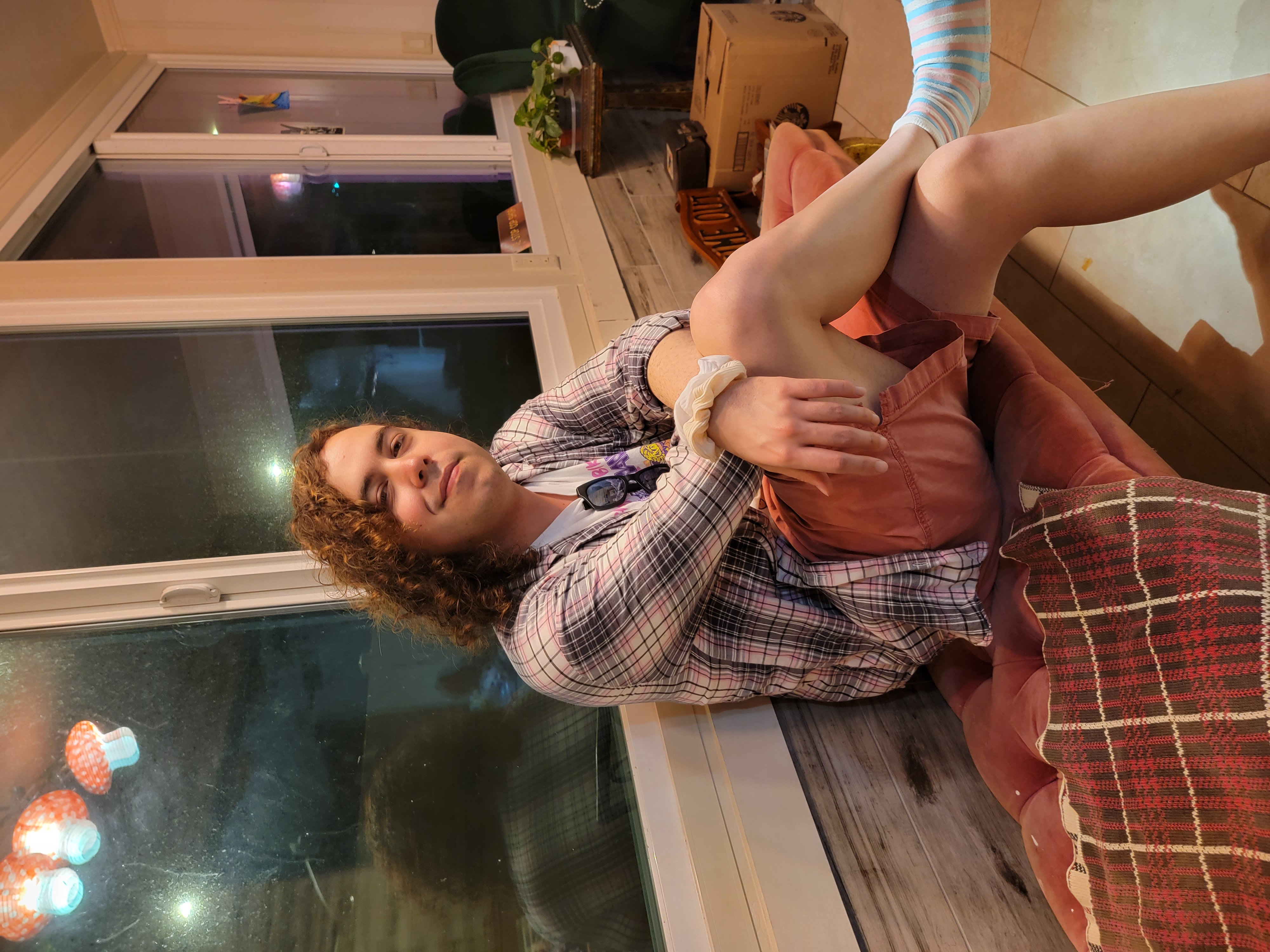 Kass is shown seated cross-legged on a cushioned bench with windows behind her, facing the camera at a three-quarter angle. She is wearing orange shorts, a grey plaid button down, unbuttoned with a white t shirt underneath it. Her brown curly hair is loose over her shoulder, with a white scrunchie on her wrist closest to the camera.