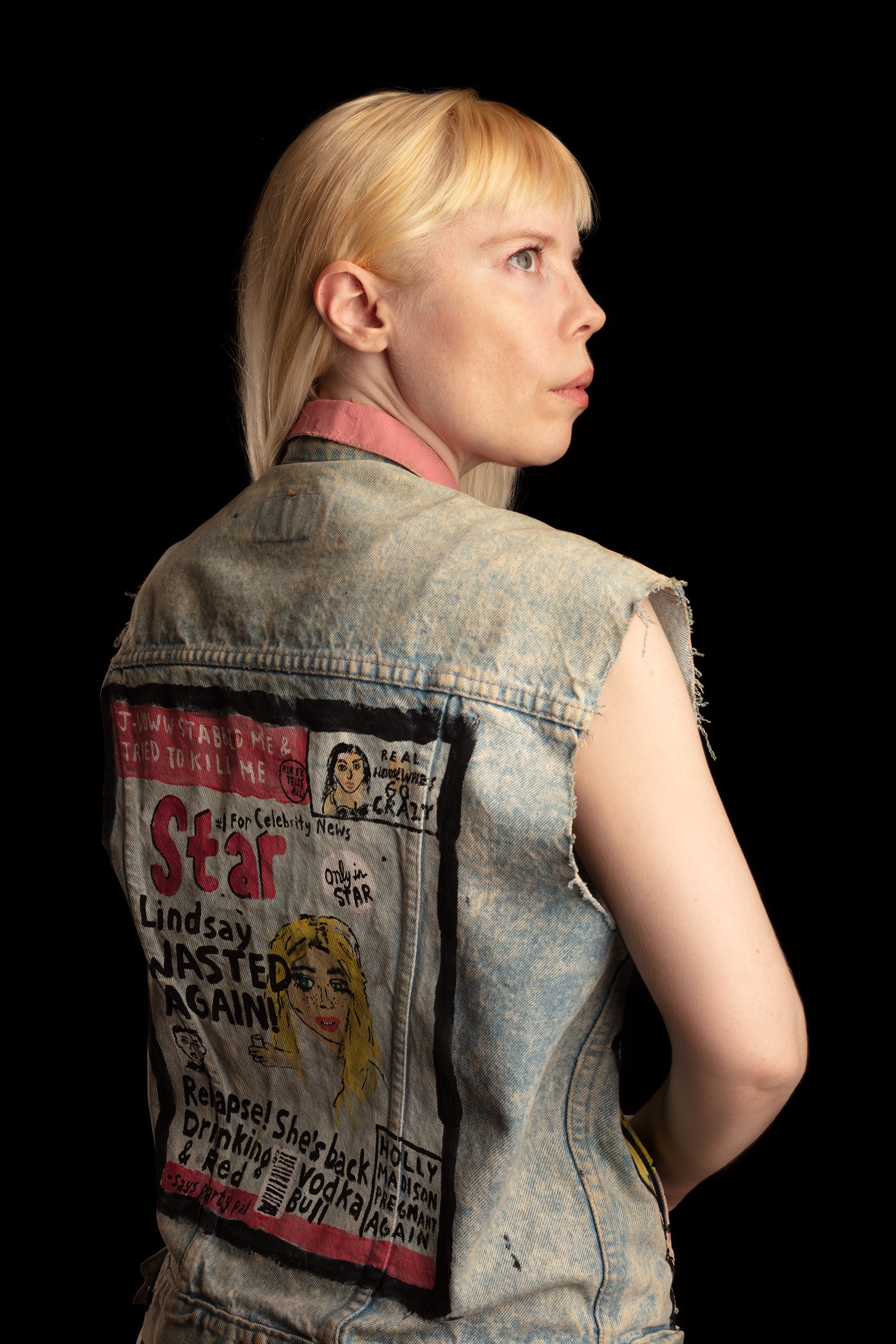 Kate Durbin is standing facing away from the camera, with her face visible in profile, wearing a cutoff denim vest with an illustrated tabloid cover on the back.