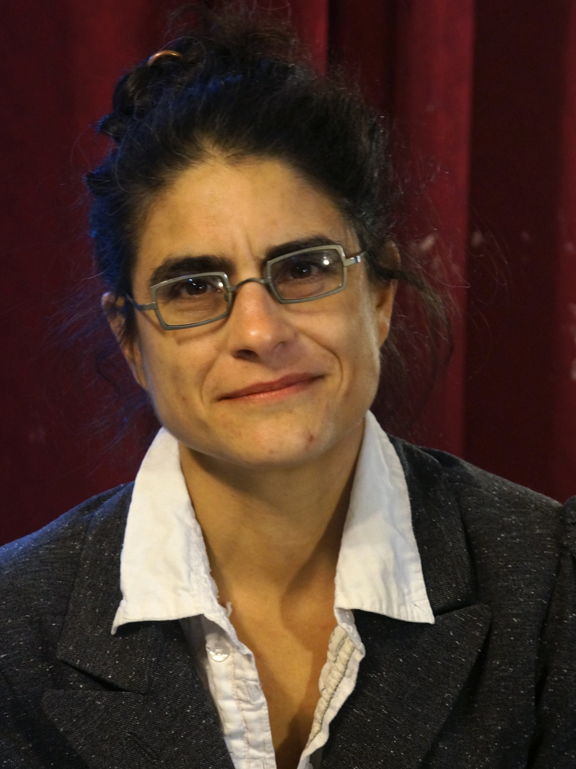 Head and shoulders of a smiling white woman with dark hair in a bun, in a white collared shirt, a gray jacket, and square glasses.