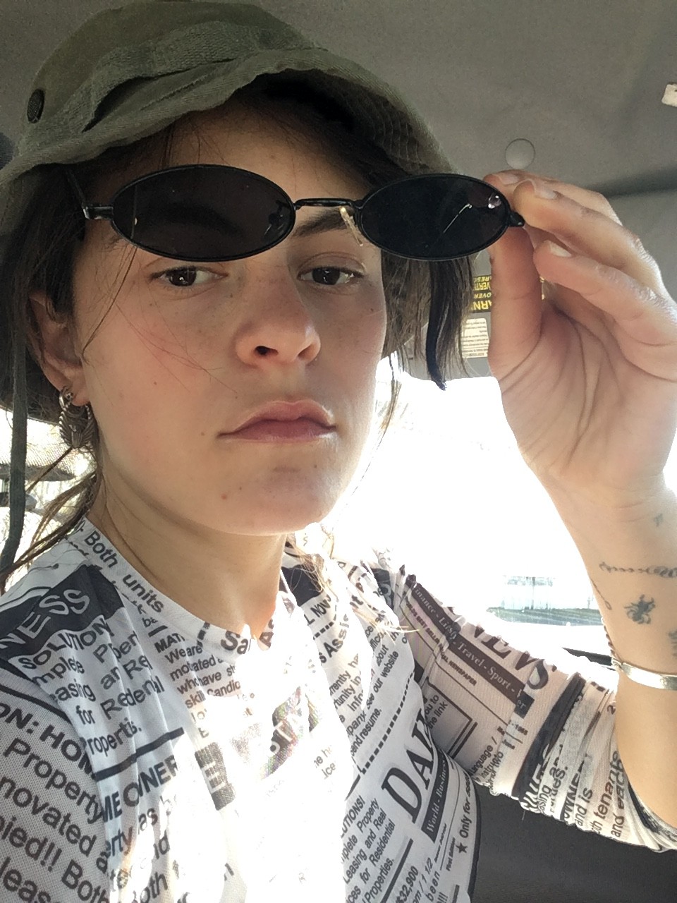 Kayla Ephros is sitting in a car, wearing a shirt with newsprint design on it, lifting her sunglasses up to look at the camera