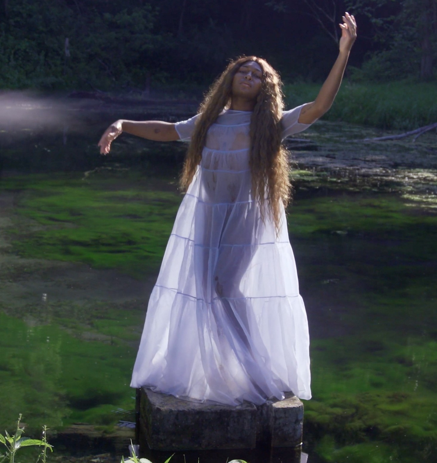 In this film still from Christopher Sonny Martinez's Come Hell or High Femmes (2021), Keioui Keijaun Thomas stands with her long hair down on a stone platform over moss, wearing a floor length gauzy dress, with one arm extended above her head and her other extended to the side.