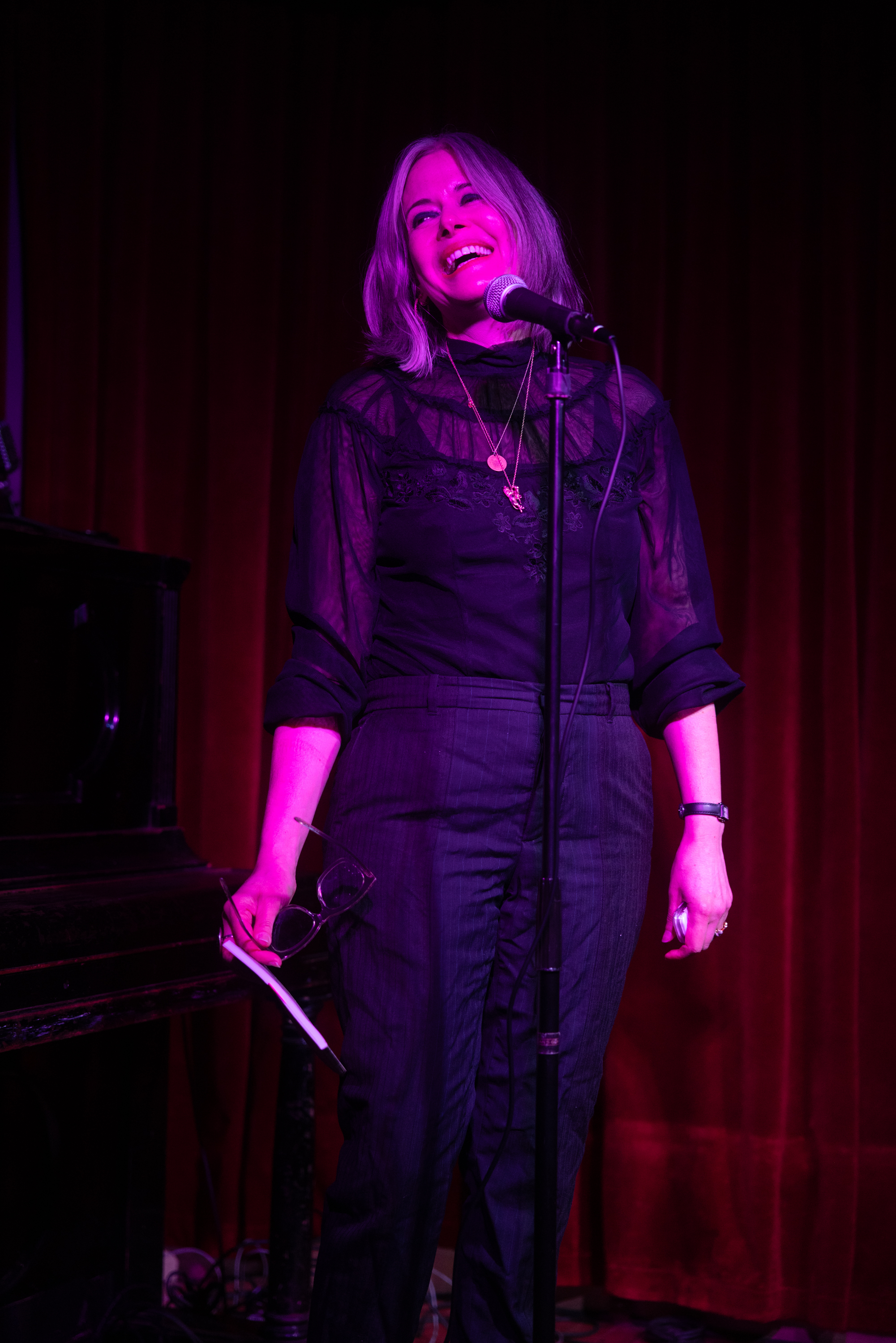 Kim Rosenfield, a white woman with straight, shoulder-length steel gray hair, is onstage at a dark club, under purple stage lights, standing at a podium with a microphone.  She has on a high-necked black lace blouse,  two gold necklaces, and black wool trousers.  She is looking to the right and is laughing.