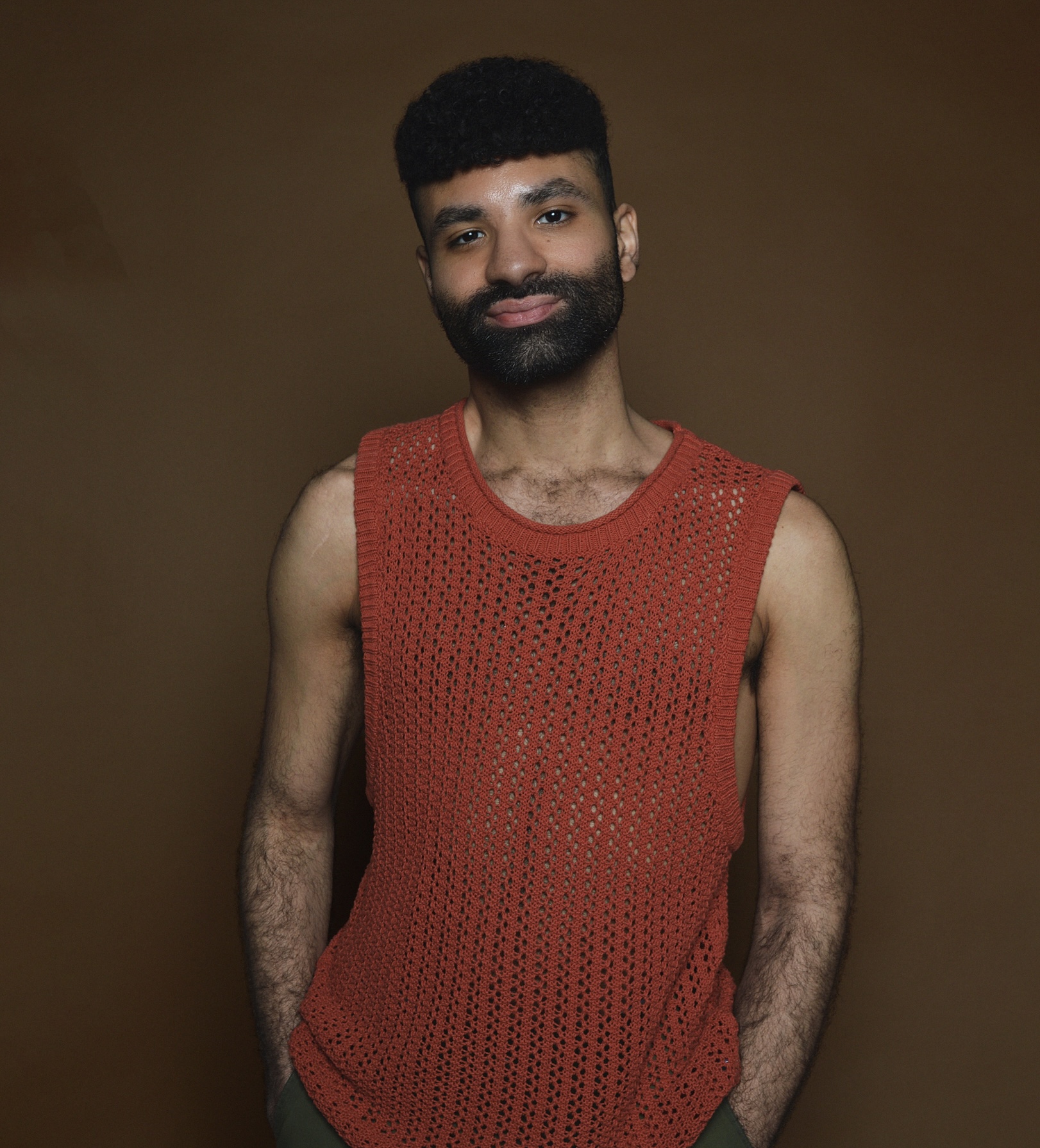 Kyle Carrero Lopez, light-skinned Black man with curly black hair and a beard wearing a copper-toned, sleeveless woven top, smiles in front of a brown background.