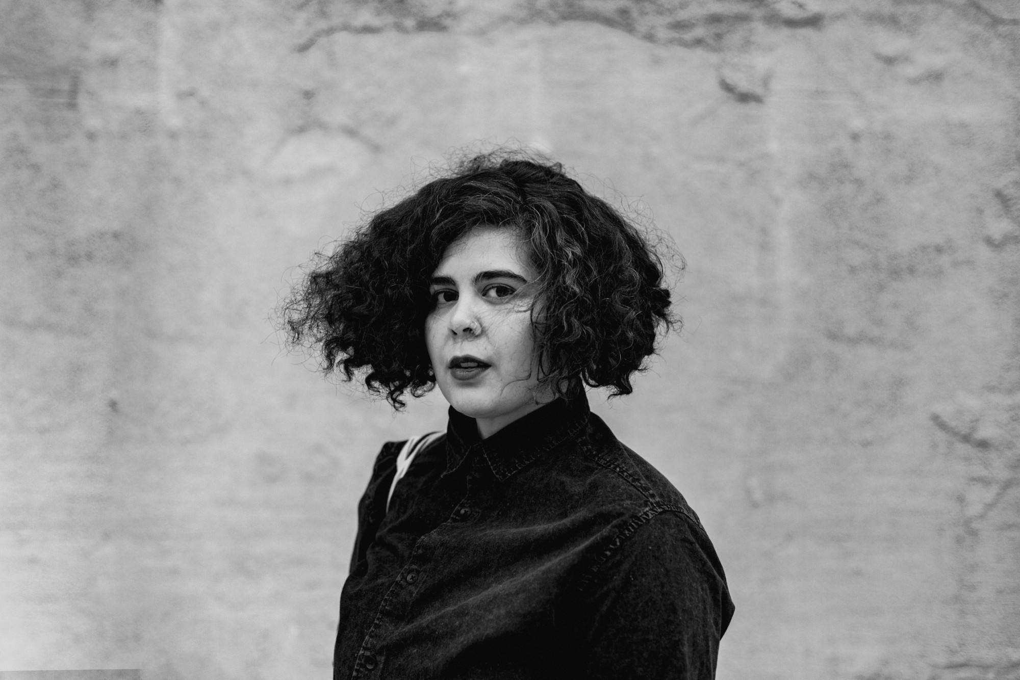 In this black and white portrait, Lara Mimosa Montes wears a dark button-up shirt and stands in front of a wall that looks like concrete, looking into the camera at an angle