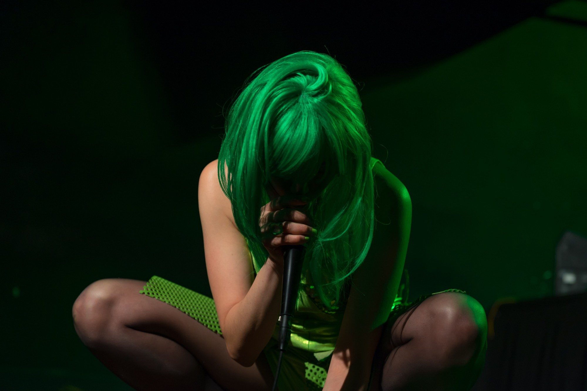 Leyya Mona Tawil [Lime Rickey International] is seated and crouched onstage, centered in a dark photograph. She is hunched over, holding a microphone. Her face is obscured by shadows and a lime green wig.