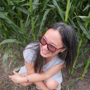 Lily Hoang is photographed from above, sitting on the ground next to what appears to be cornstalks, looking to the side and smiling