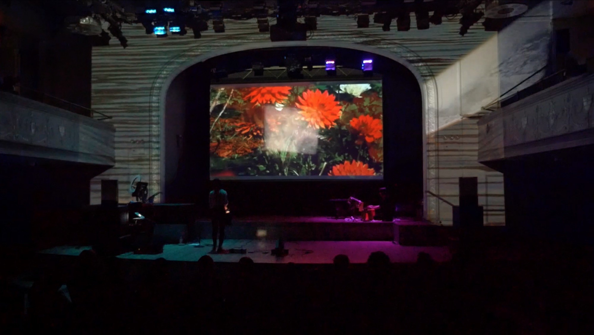 Photo of a performance where two performers occupy opposite sides of a stage, in front of a projection of red flowers